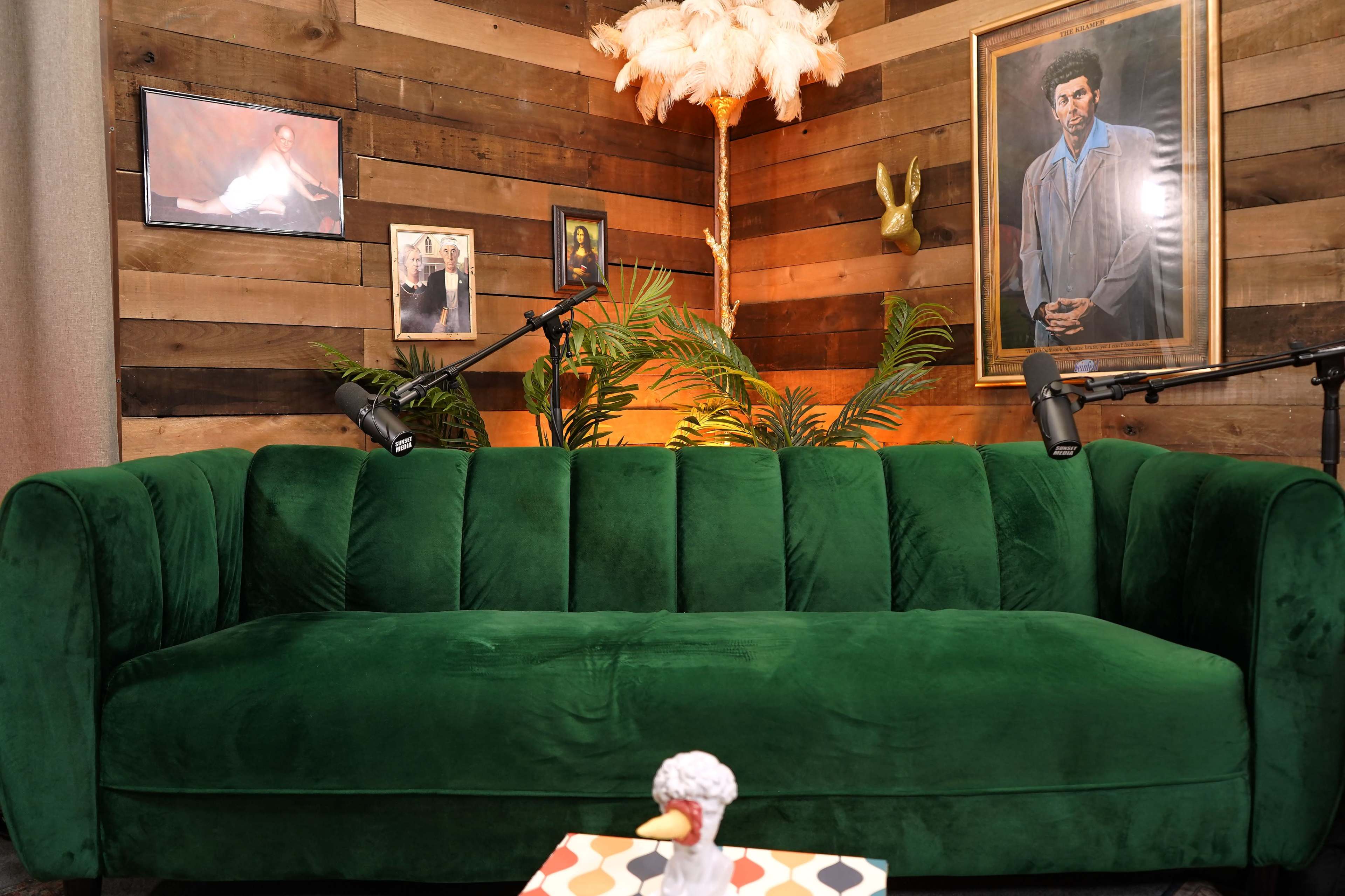 The image shows a green velvet couch positioned in a rustic room adorned with wooden walls, framed portraits, and decorative plants, illuminated by warm lighting.