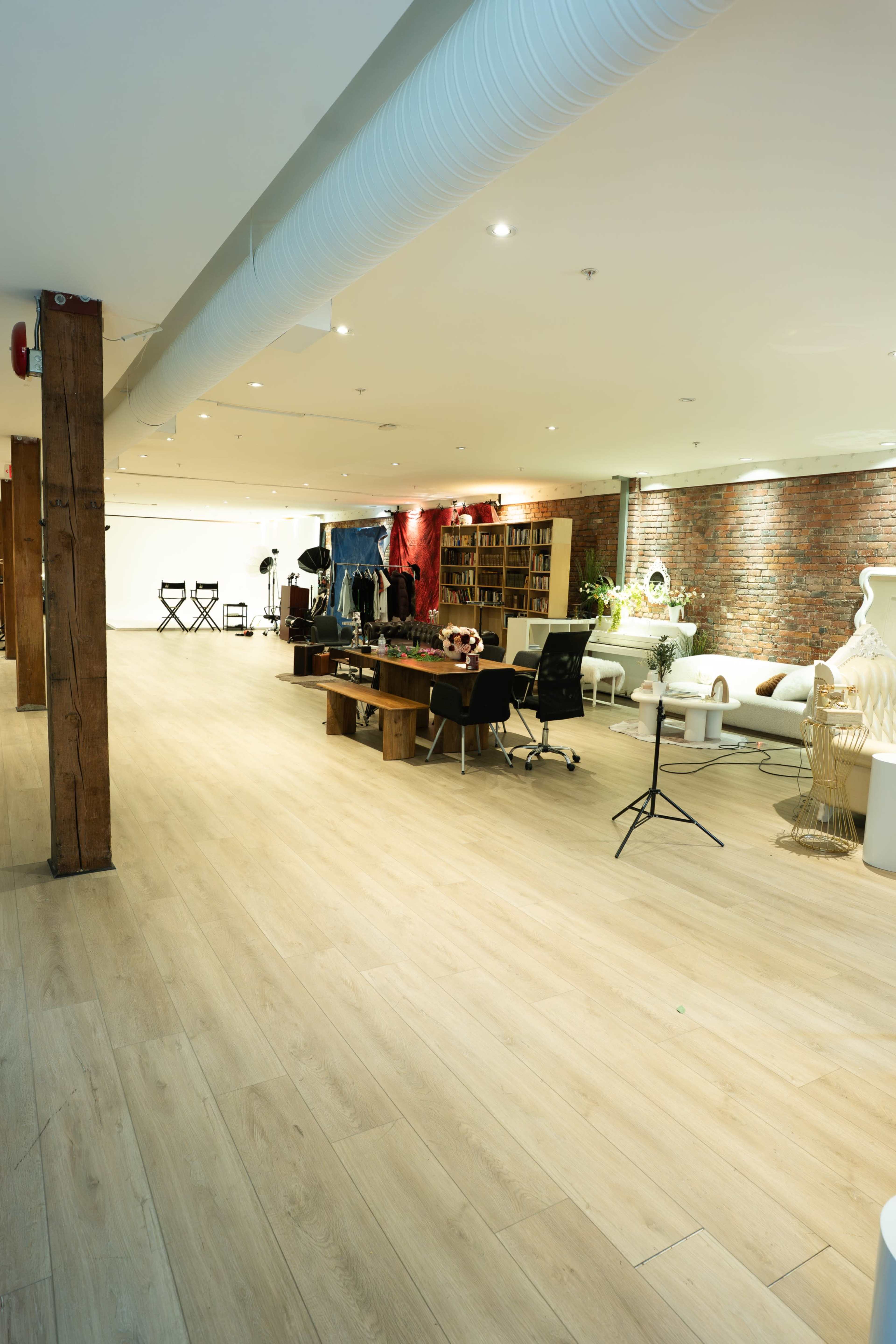 The image shows a spacious interior featuring wooden flooring, exposed brick walls, and a mix of furniture, including a long table, chairs, and shelves filled with books.