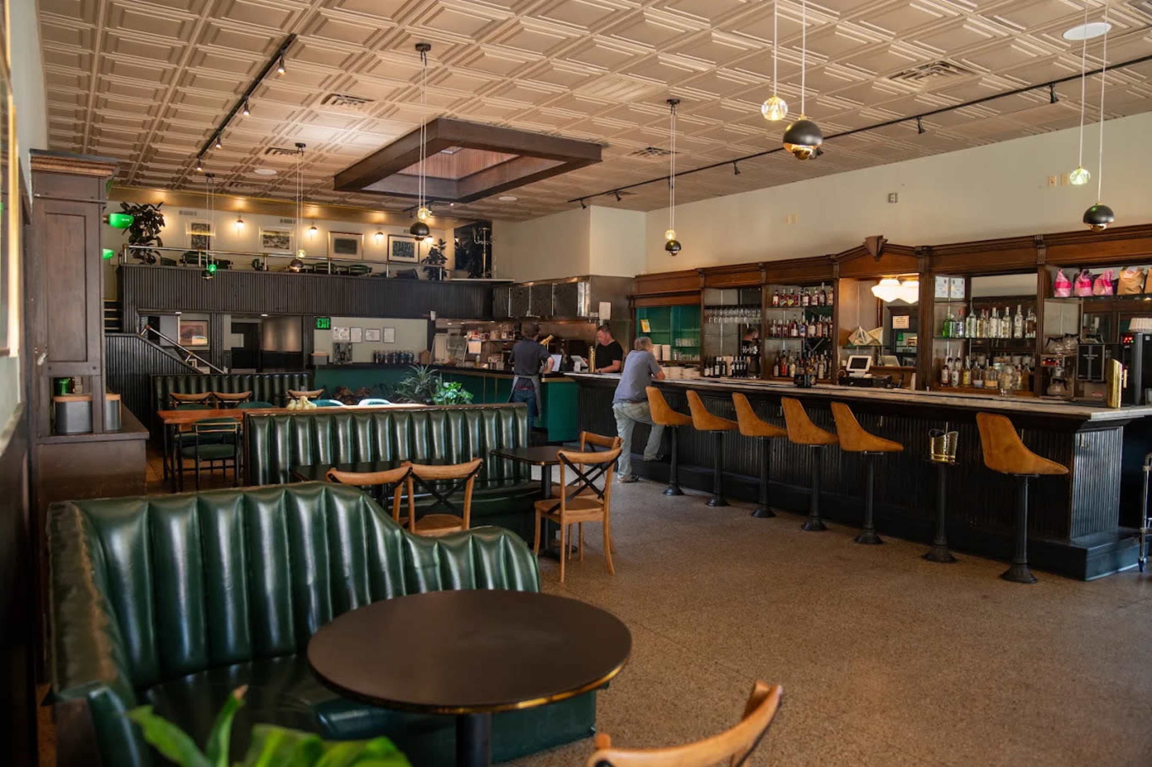 The image shows a spacious, vintage-style bar with green leather seating, wooden furniture, and a long counter where two people are engaged in conversation.