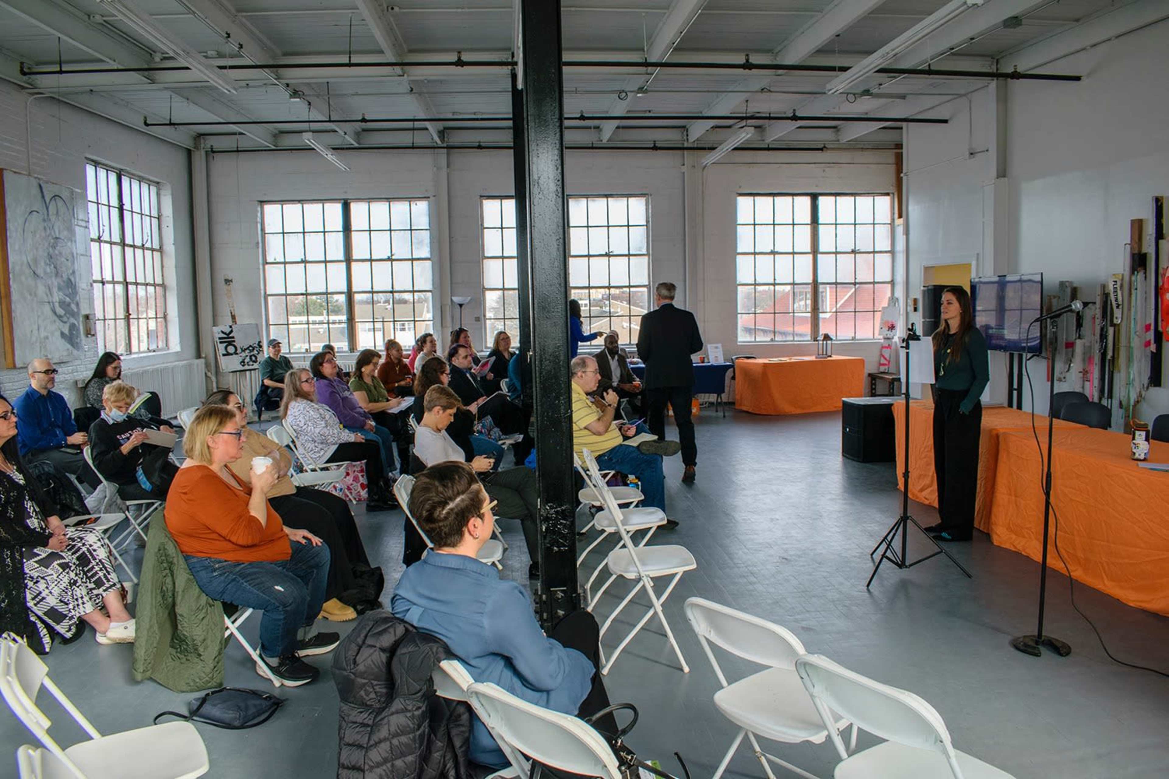 A group of people sits in white chairs arranged in a spacious room with large windows, while a speaker stands at a microphone near an orange table.