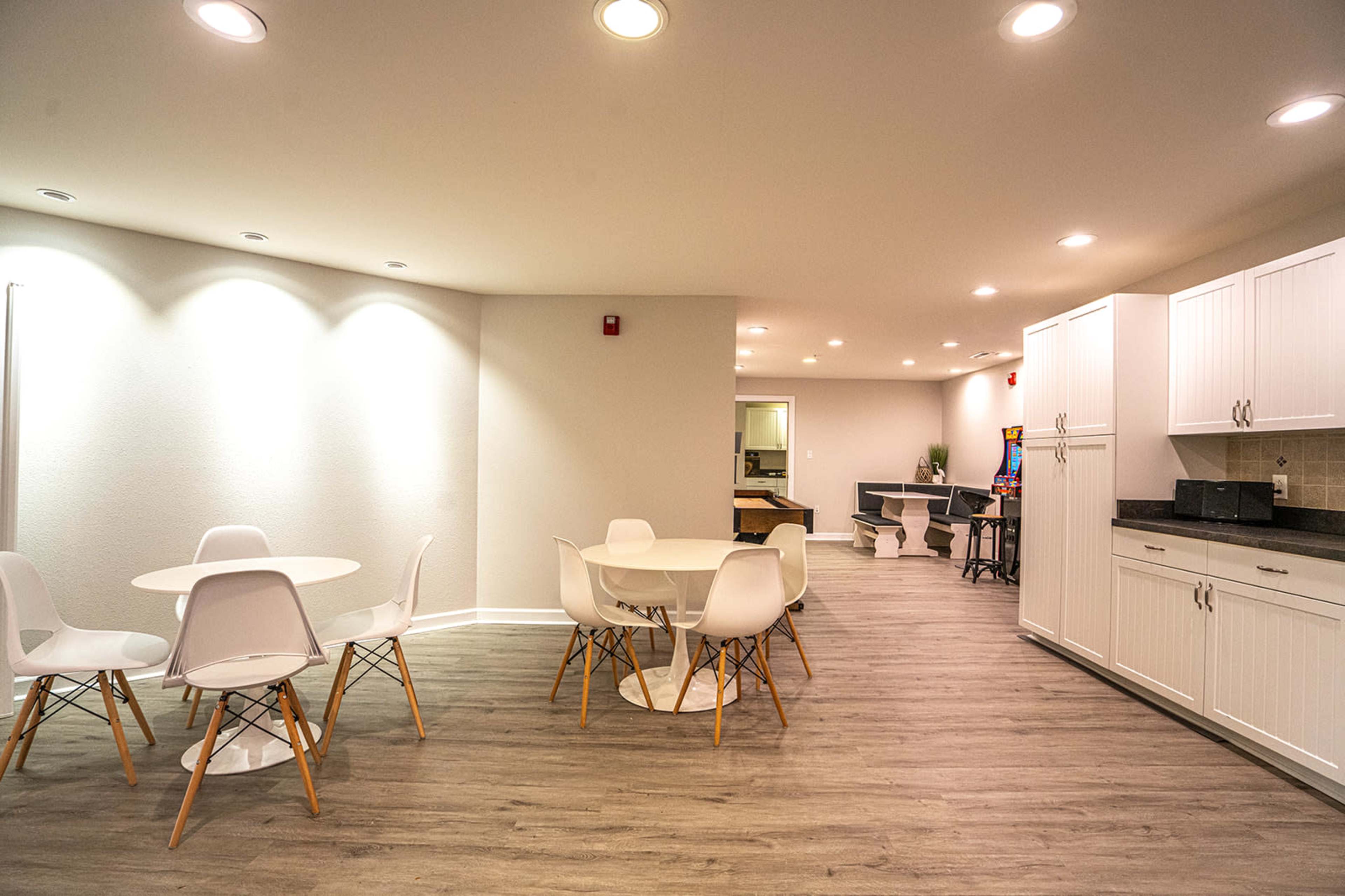 A modern, spacious interior featuring a round table with white chairs, a kitchenette, and an adjacent area with additional seating and workspaces.