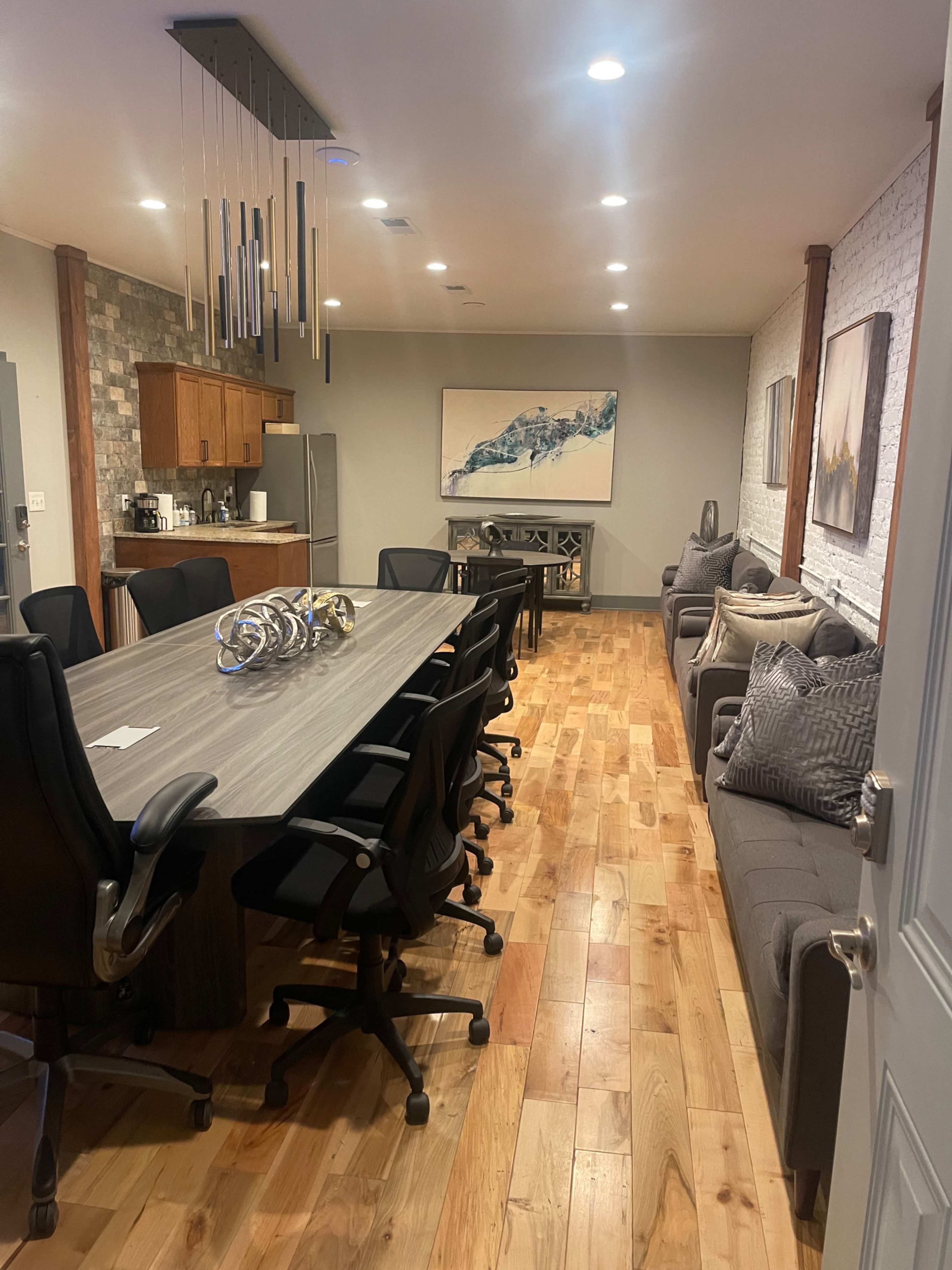 A modern office space featuring a long conference table surrounded by black chairs, with a kitchen area and artwork on the walls.