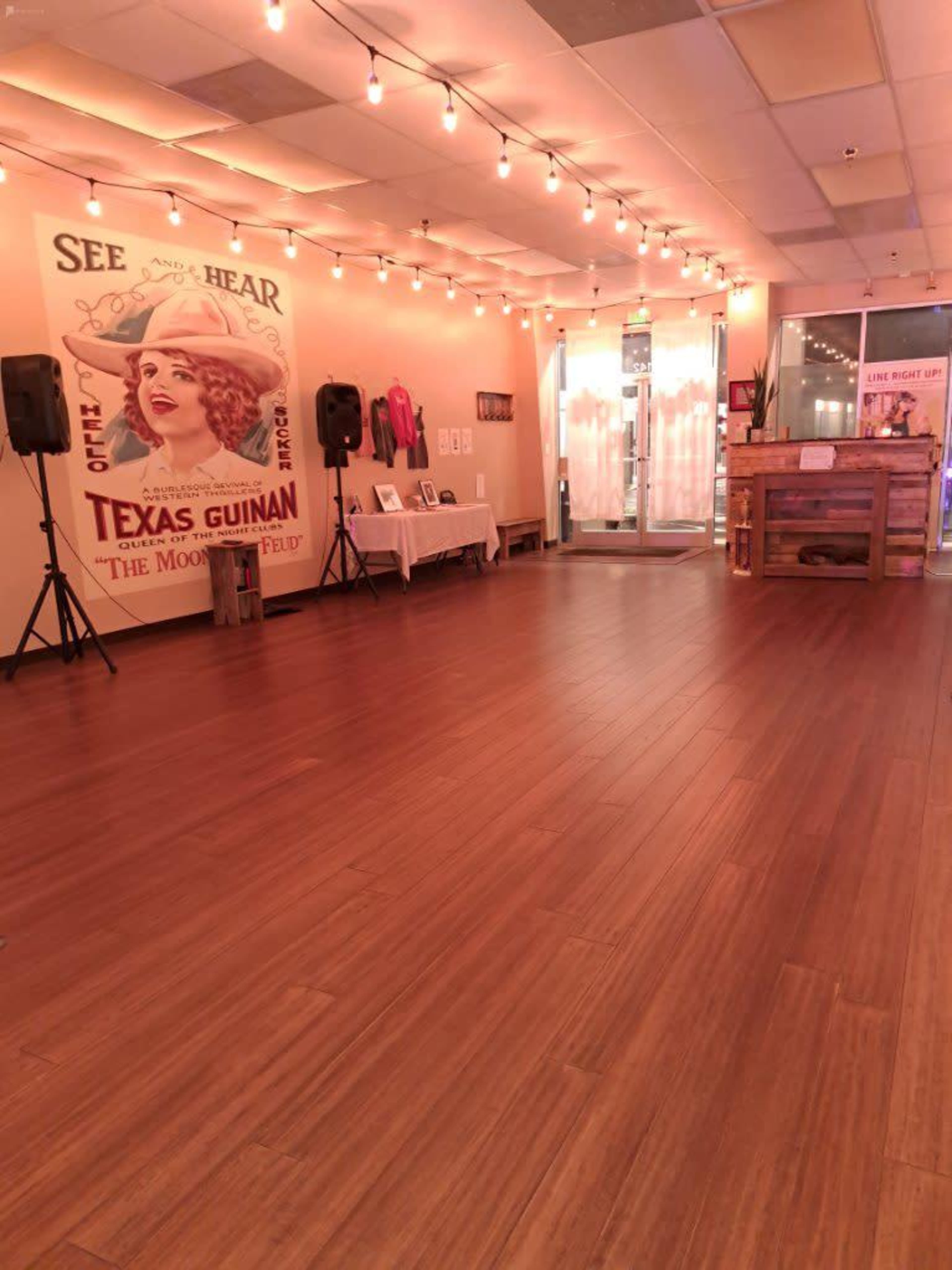 A spacious room with wooden flooring, string lights overhead, a decorative poster on the wall, and a reception area with a wooden counter.