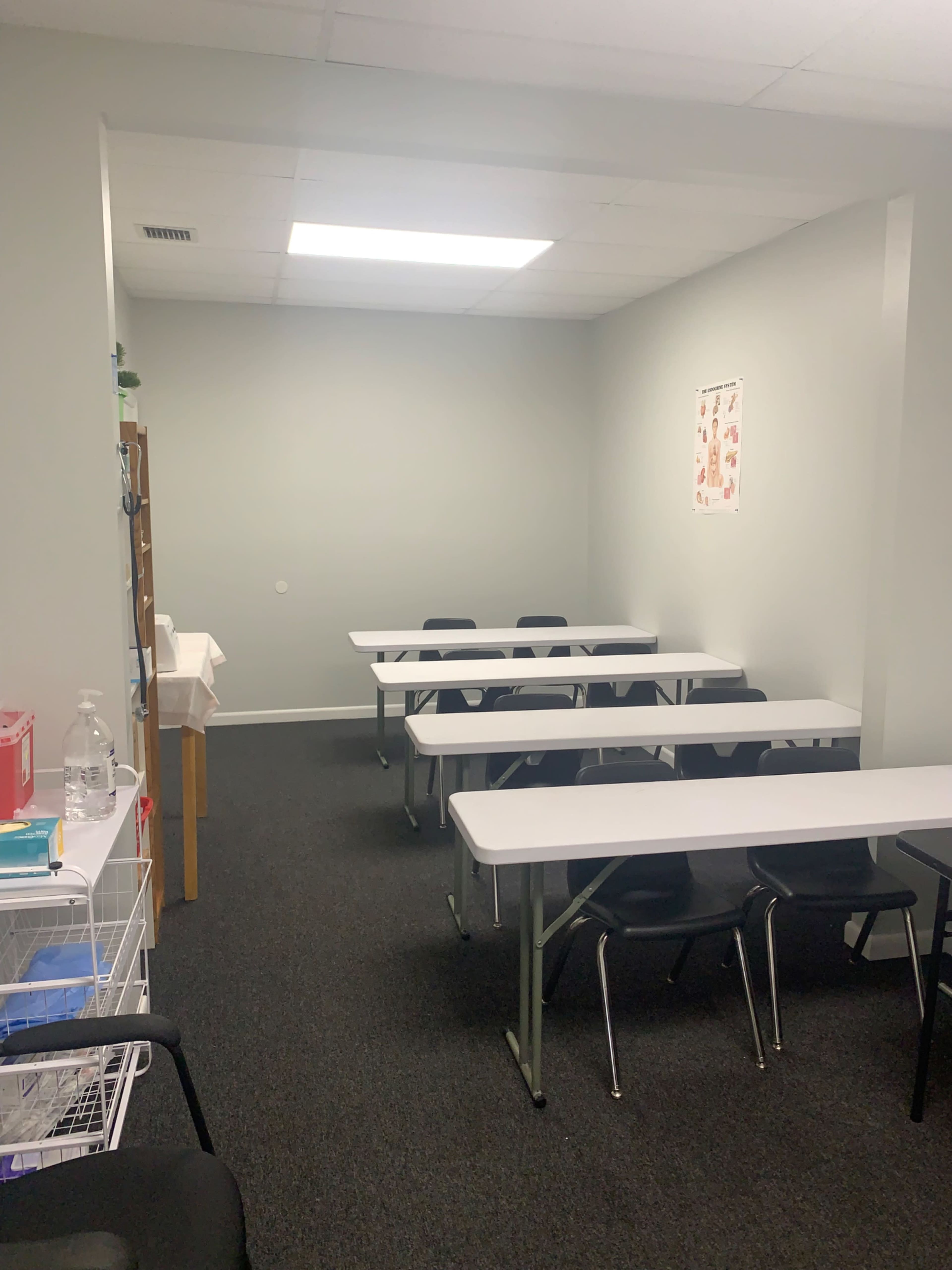 A small, empty classroom with two sets of white tables and chairs arranged neatly on a dark carpet.