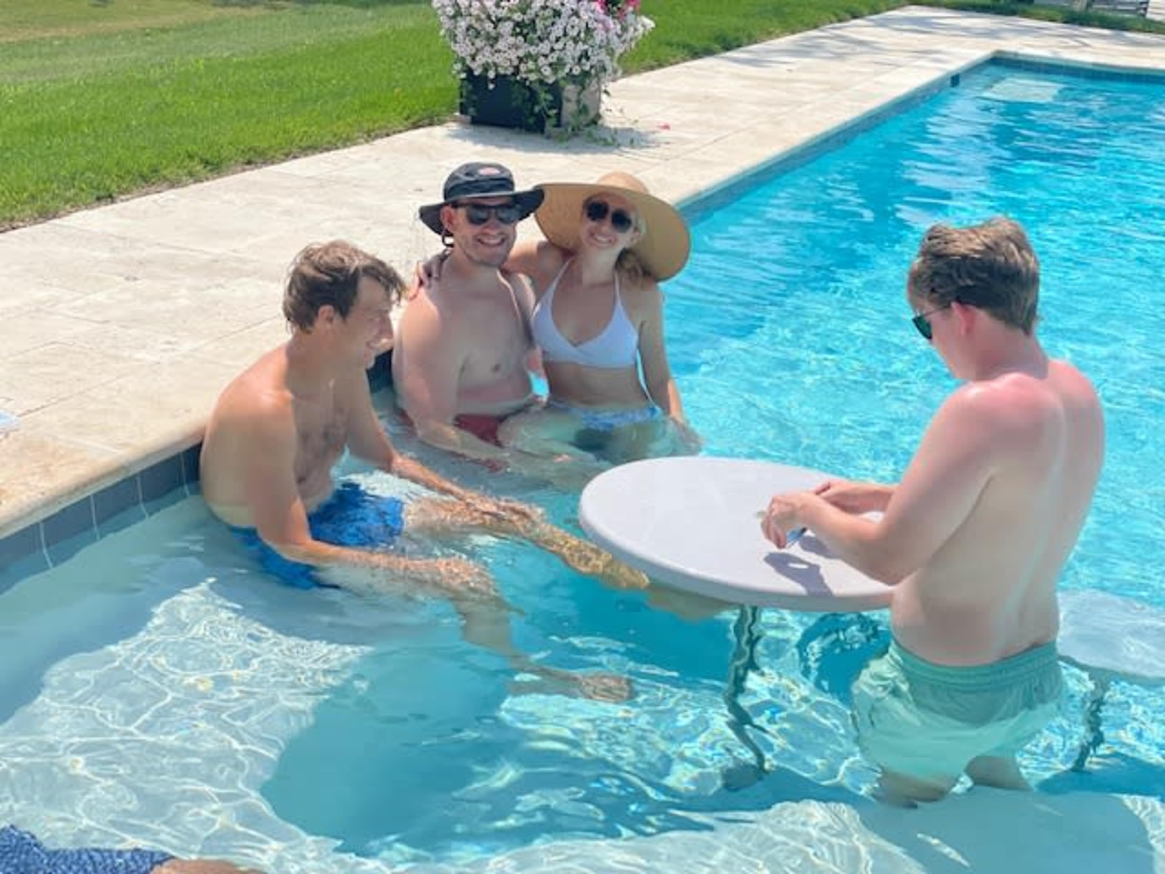 A group of four people relax in a swimming pool, with two sitting on steps and two standing nearby, while a table floats in the water.