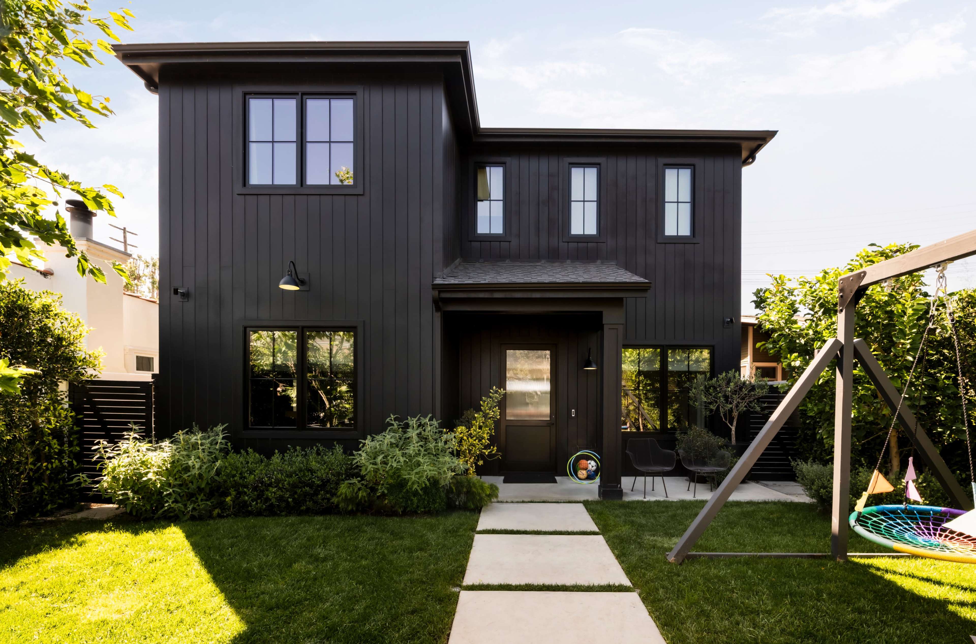 The image shows a modern black two-story house surrounded by green landscaping and a stone pathway leading to the entrance.