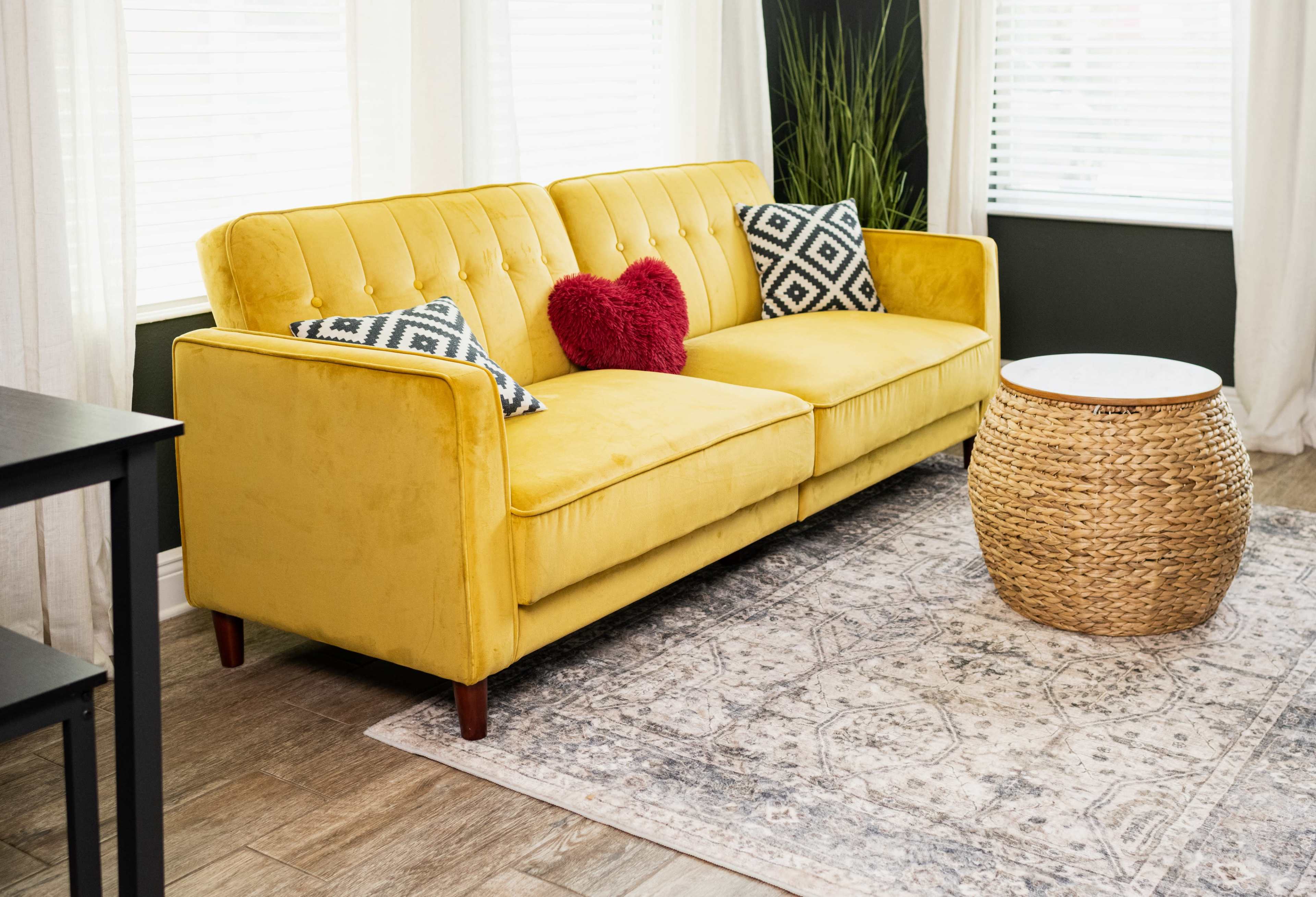 The image features a yellow sofa adorned with patterned pillows and a heart-shaped cushion, positioned on a patterned rug next to a woven side table in a well-lit room.