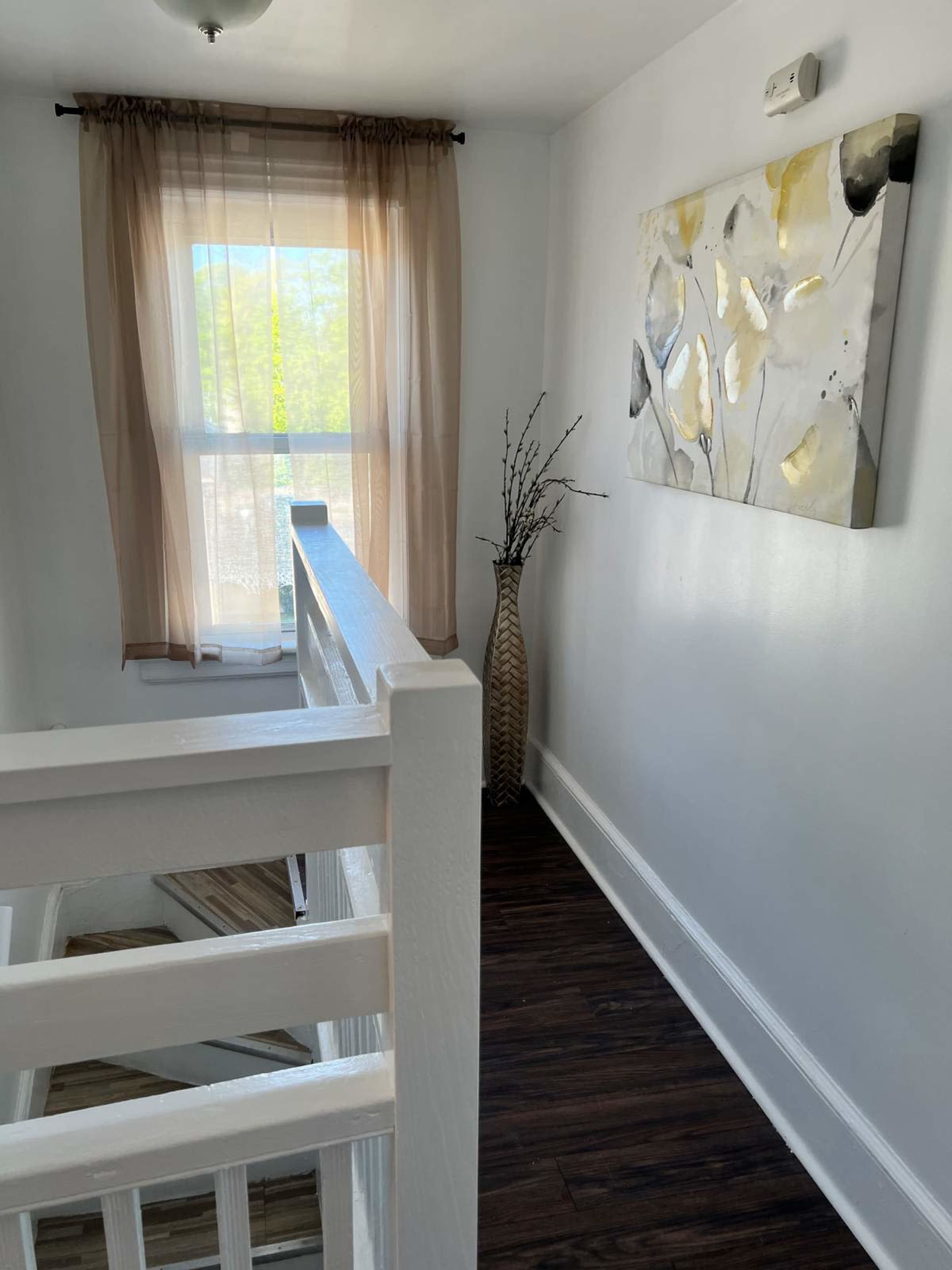 A staircase landing features a large wall art piece, a vase with branches, and a window covered with a sheer curtain.