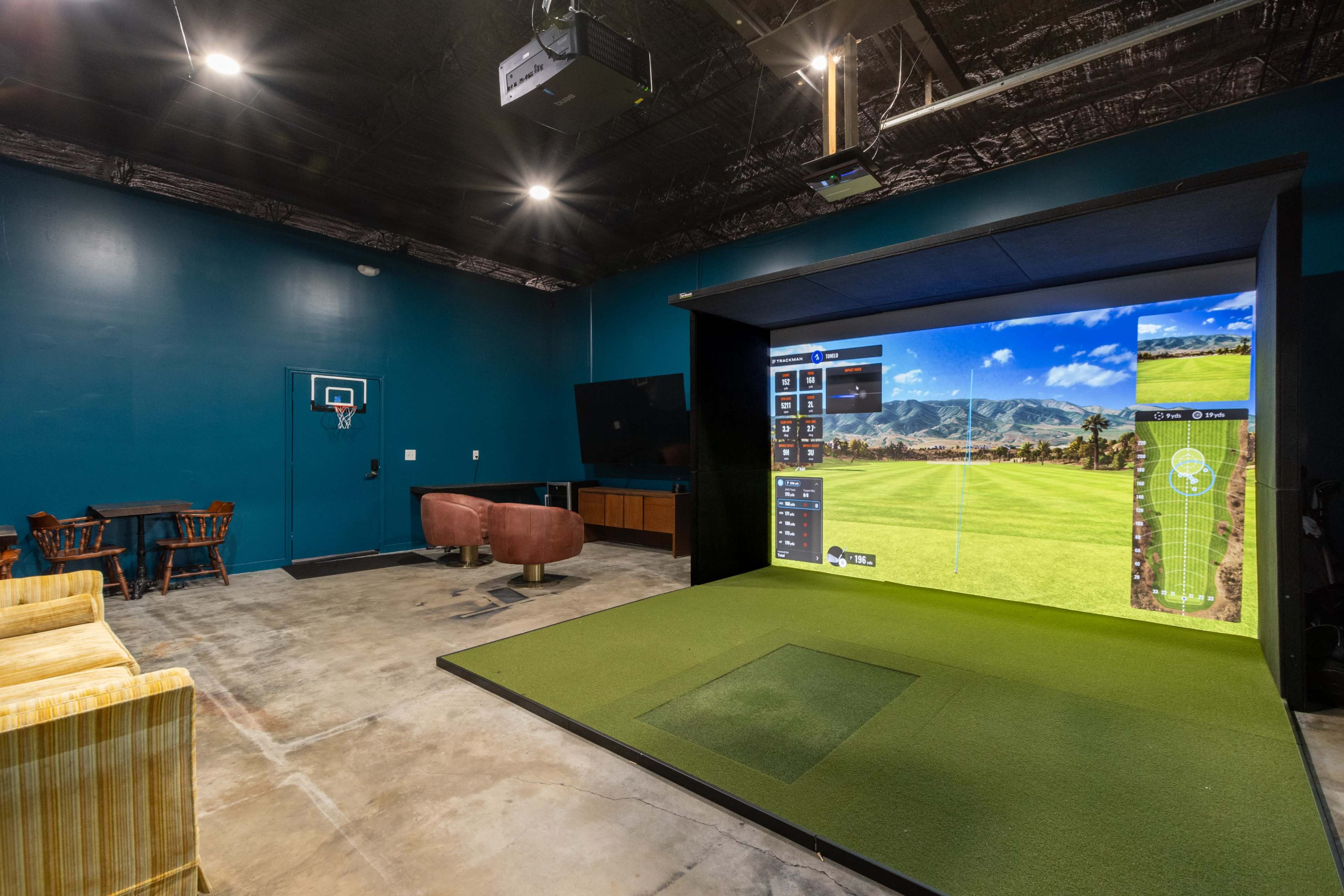 An indoor golf simulator setup with a projector screen displaying a golf course, surrounded by modern furnishings including a couch, chairs, and a small dining table.