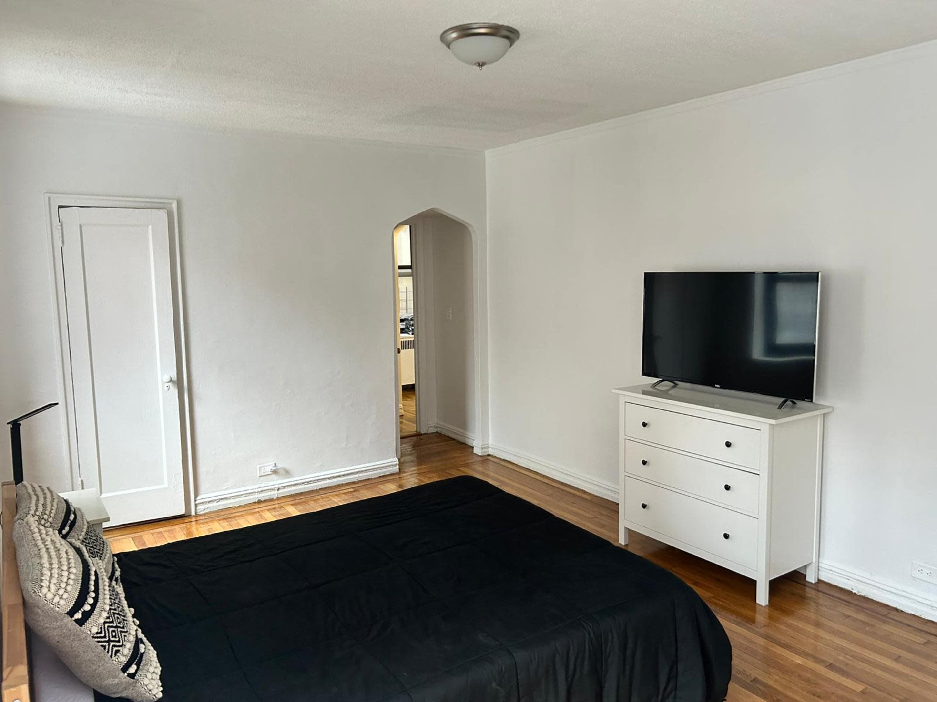 A simple bedroom features a bed with a black bedspread, a white dresser, and a television on a stand against a white wall.
