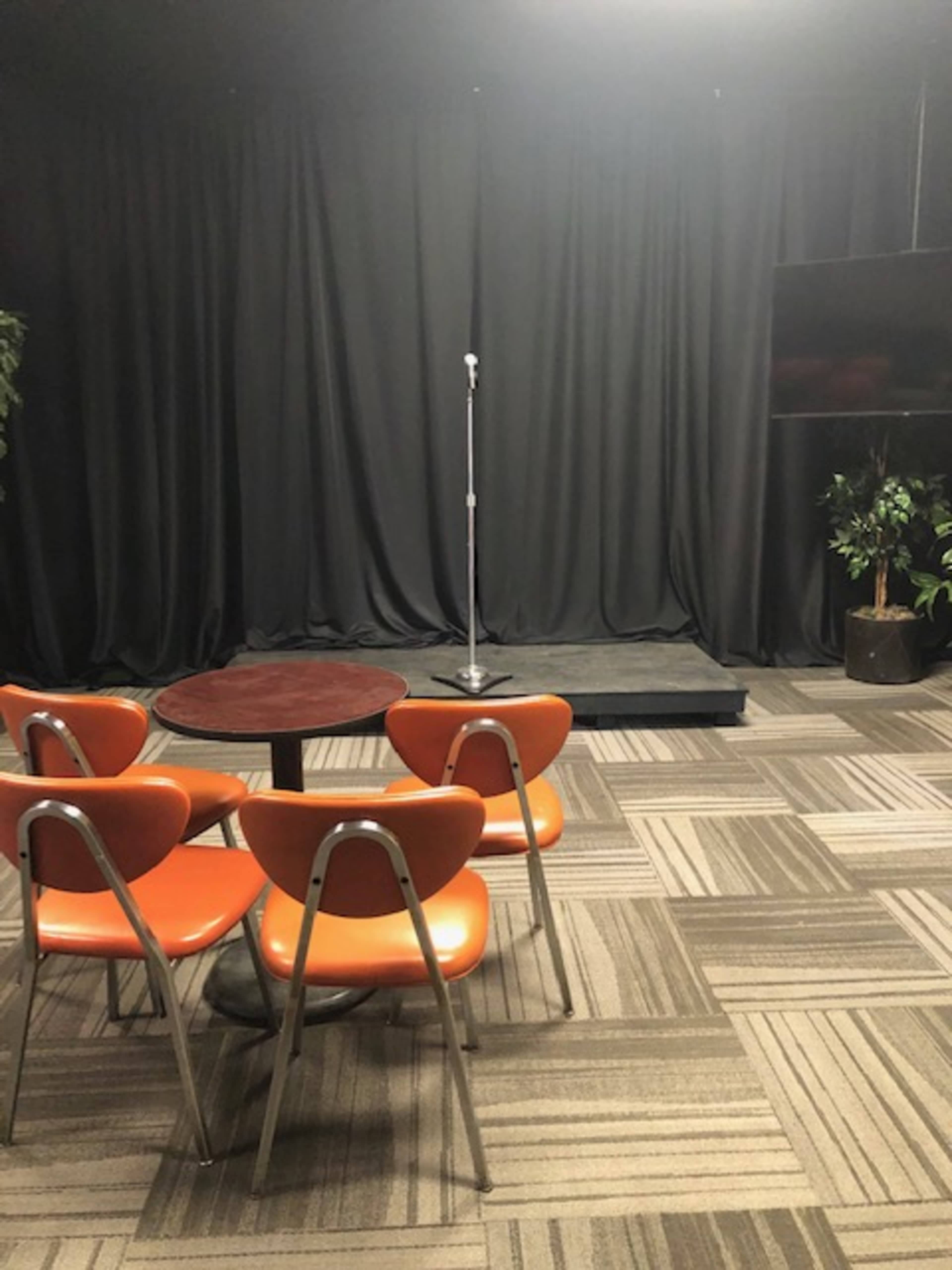 A small stage with a microphone stands in front of a dark curtain, surrounded by four orange chairs and a round table.