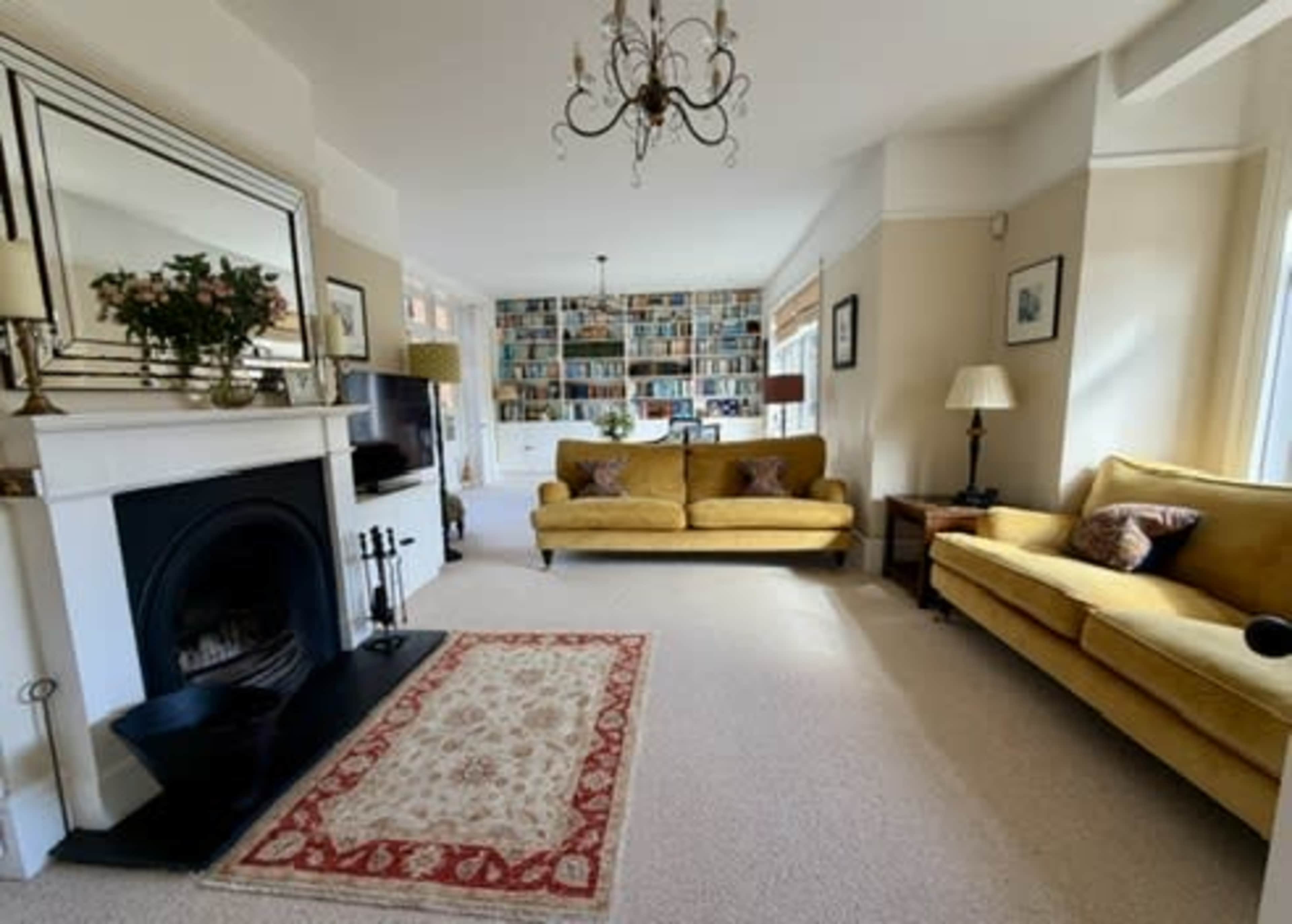 The image shows a bright living room with two yellow sofas facing a decorative fireplace, a patterned area rug on the carpeted floor, and a large bookshelf filled with books on the back wall.