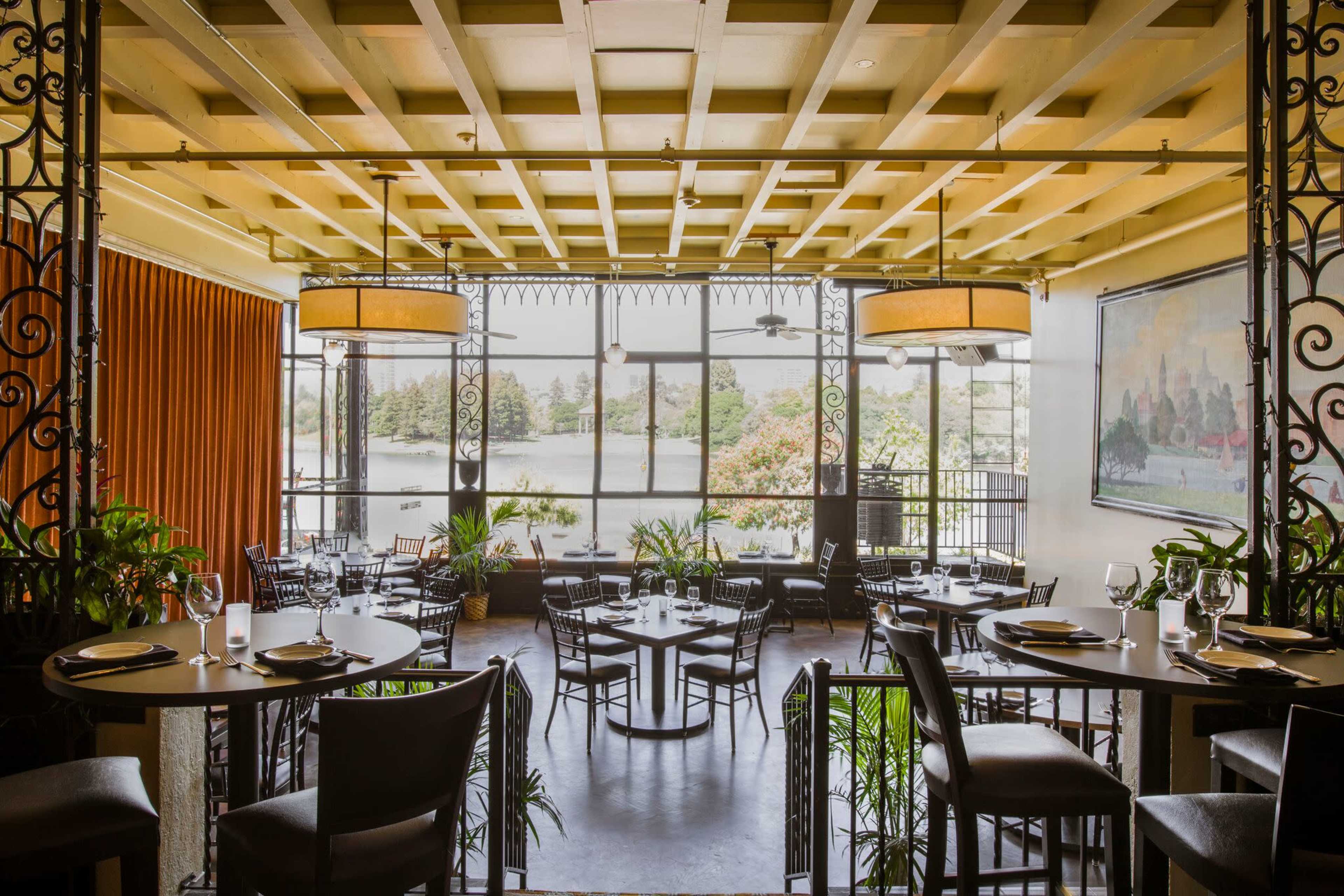 The image shows a dining area with tables and chairs arranged around a large window that provides a view of a body of water, surrounded by vibrant greenery.