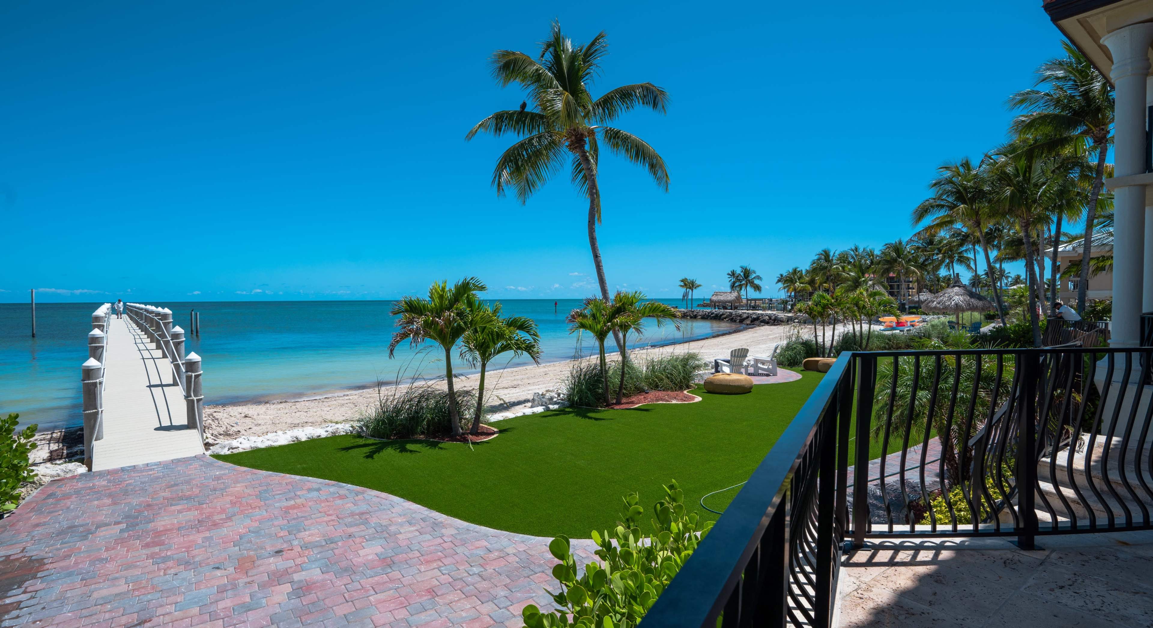 A beachfront property with a manicured lawn, palm trees, and a view of the ocean under a clear blue sky.