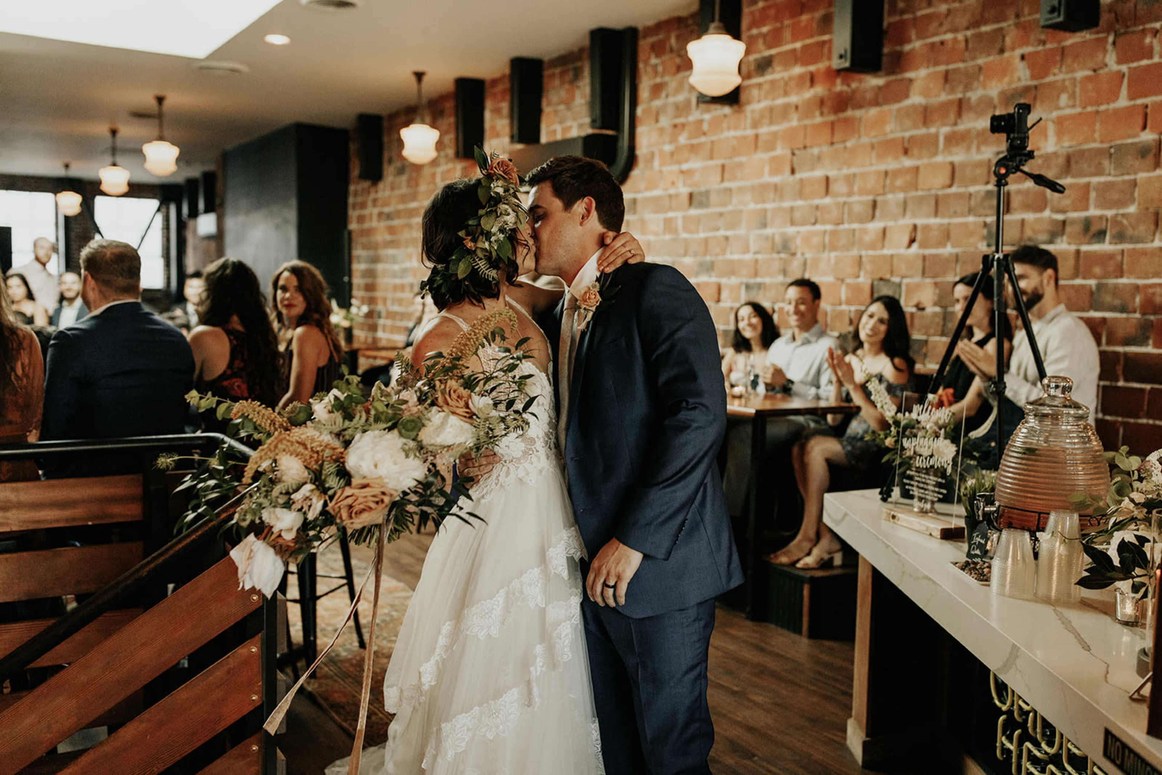 A bride and groom kiss at their wedding reception, surrounded by guests seated at tables adorned with flowers.