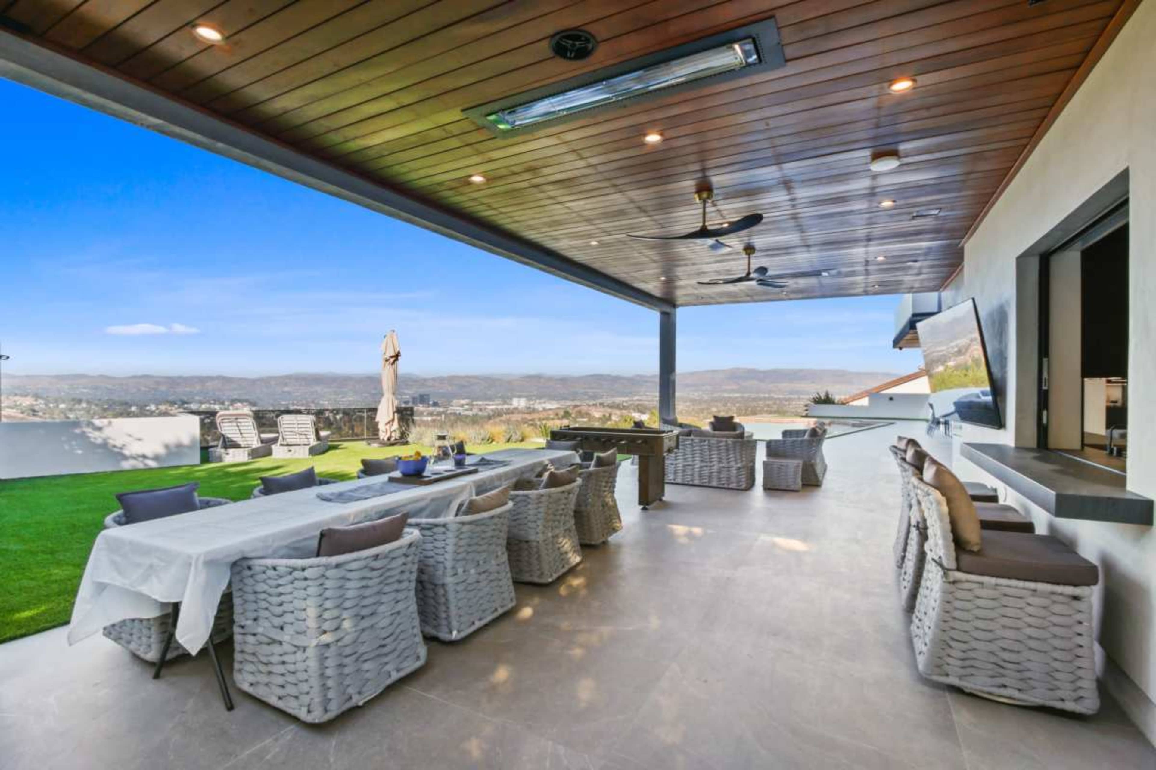 The image showcases a spacious outdoor patio area featuring wicker seating, a large dining table, and a scenic view of rolling hills under a clear blue sky.