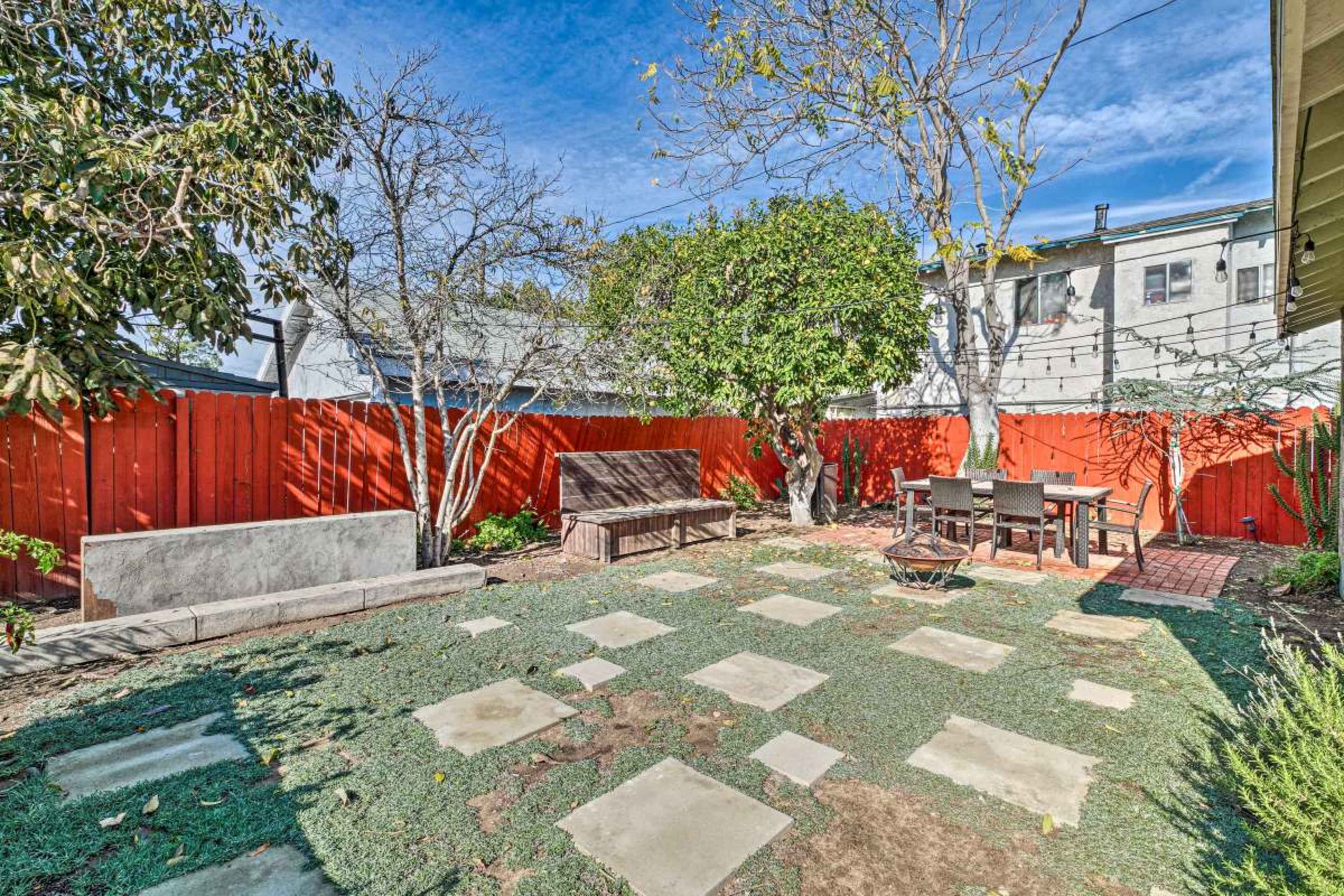 The backyard features a paved area with concrete slabs, a wooden seating arrangement, and a fire pit, all enclosed by a red fence and surrounded by trees.