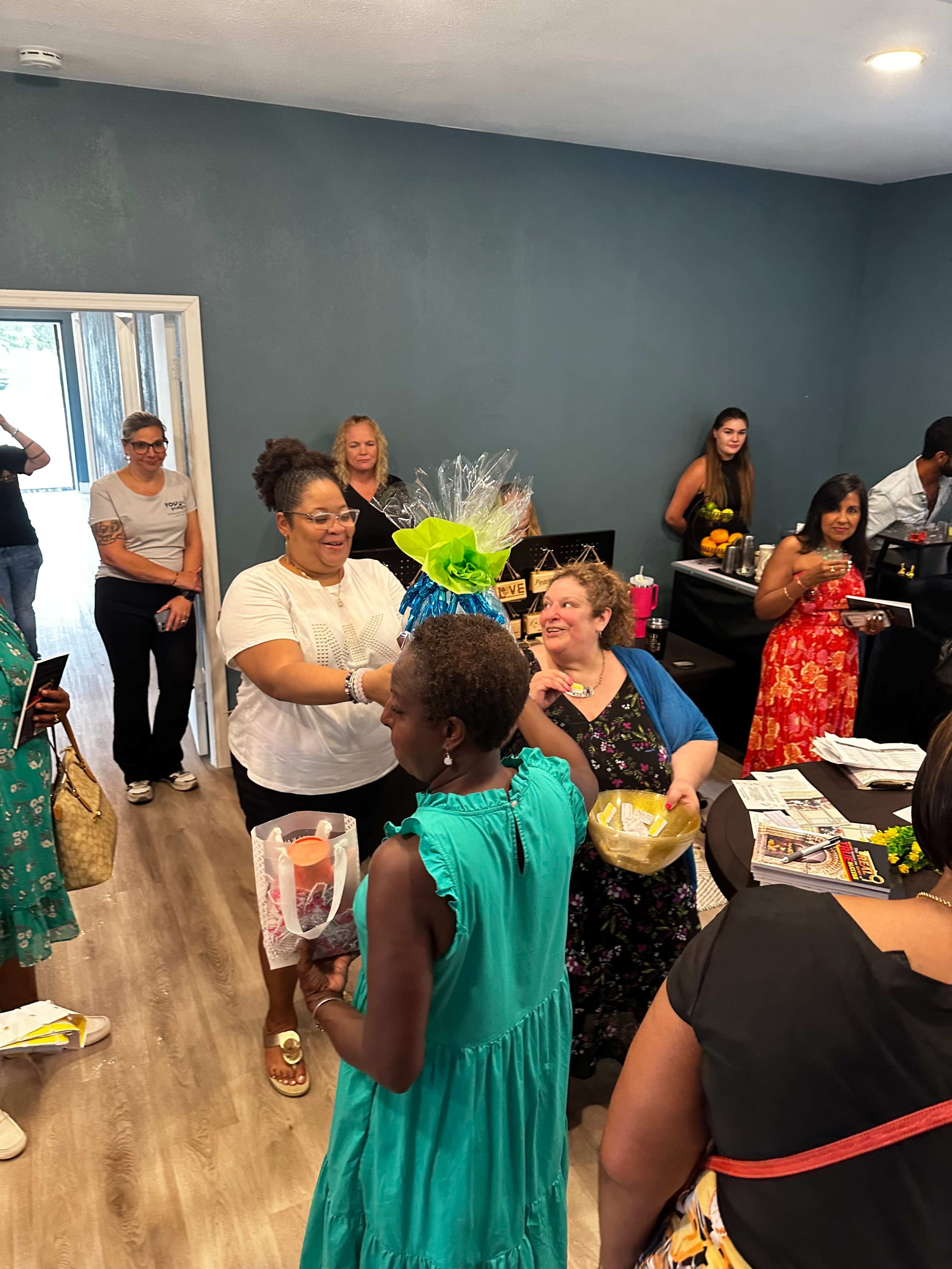 A group of people gathers in a room, engaging in conversation and celebrating with brightly colored decorations and gifts.