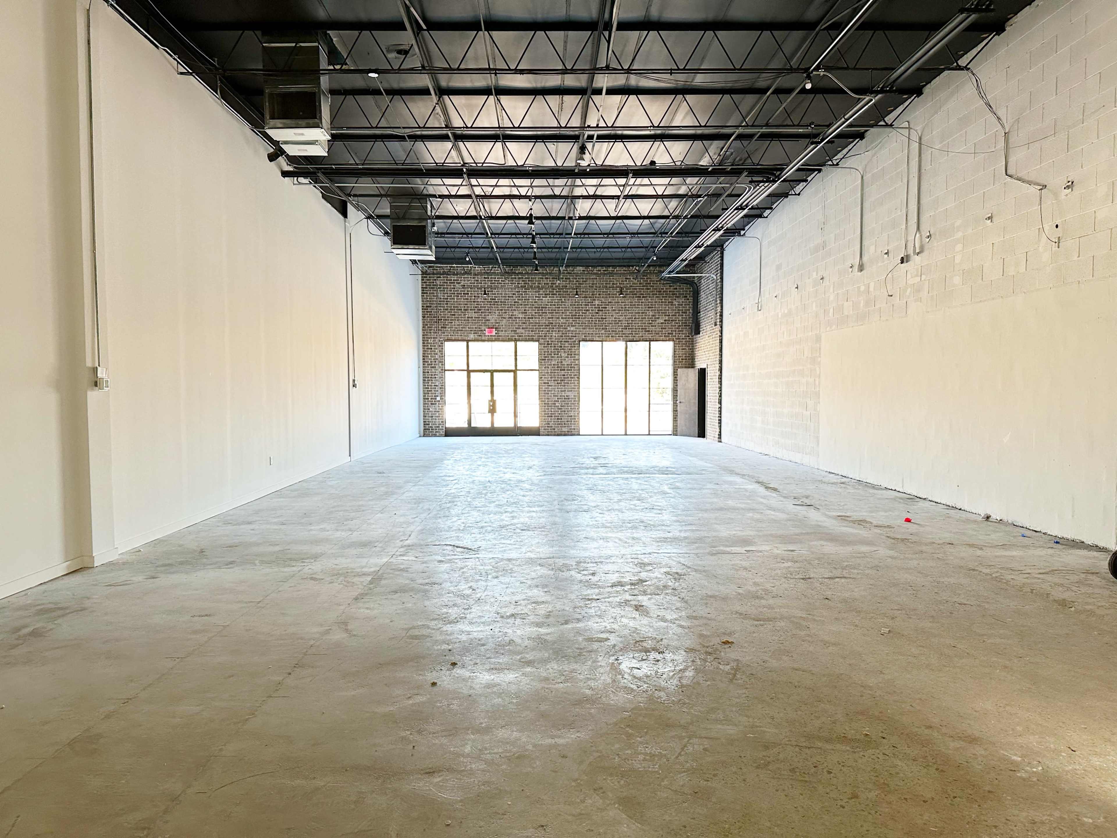 The image shows a spacious interior with bare concrete flooring and large windows at the far end, providing natural light.