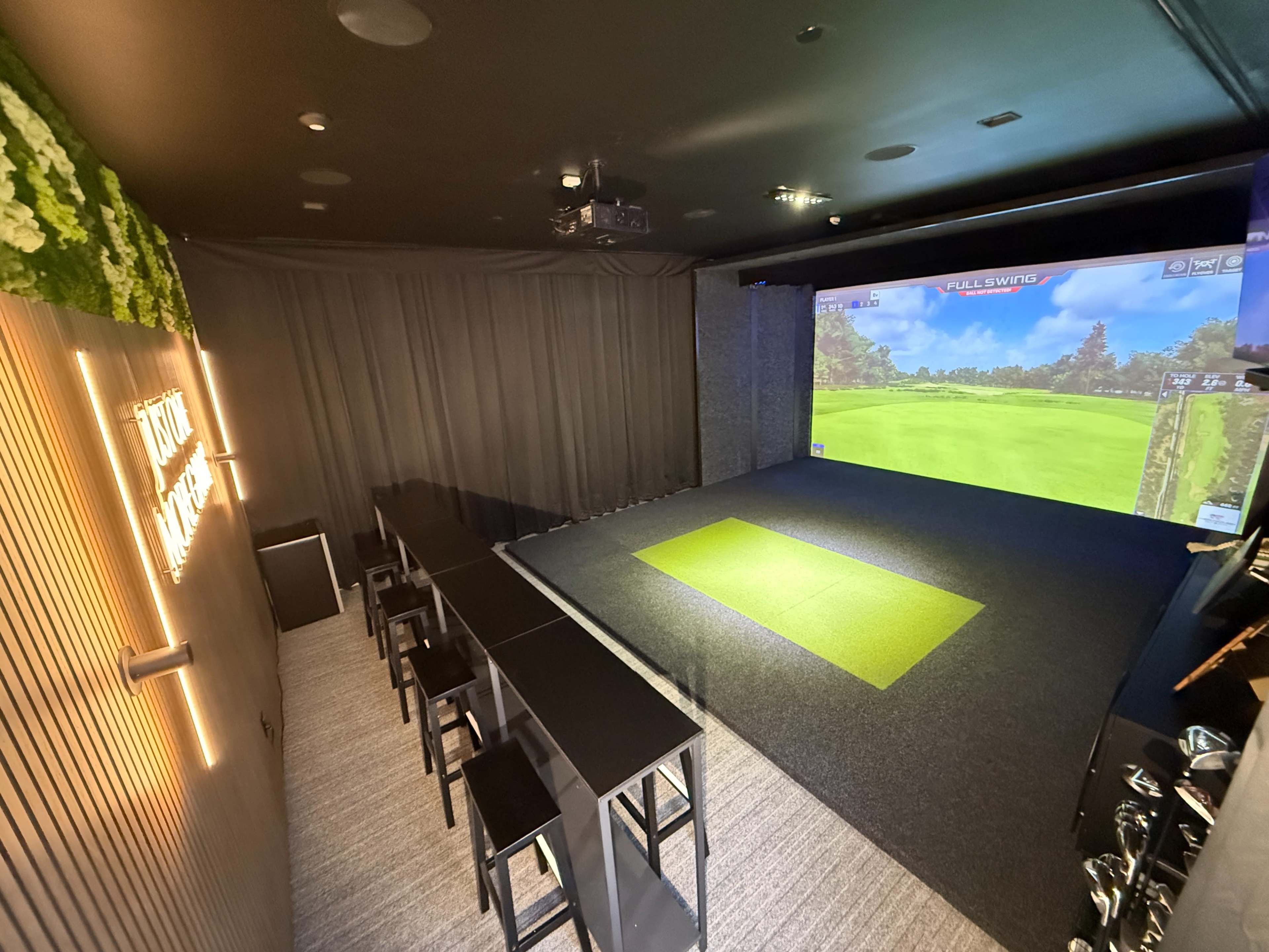 The image shows an indoor golf simulator room with a large projection screen displaying a golf course, complemented by a seating area featuring black bar stools and a long table.