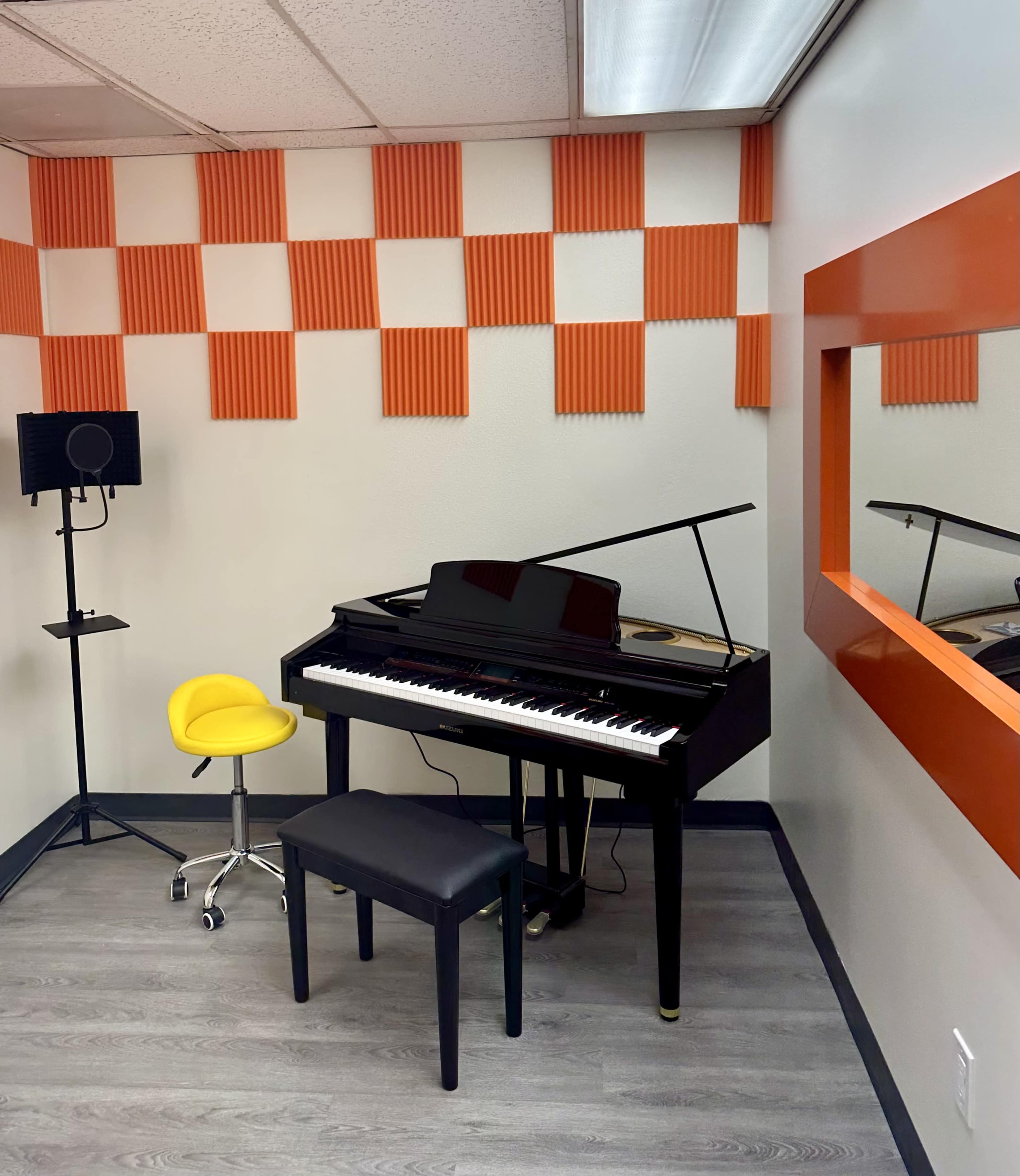 The image shows a music practice room featuring a black grand piano, a yellow chair, a black bench, and orange soundproofing panels on the walls.