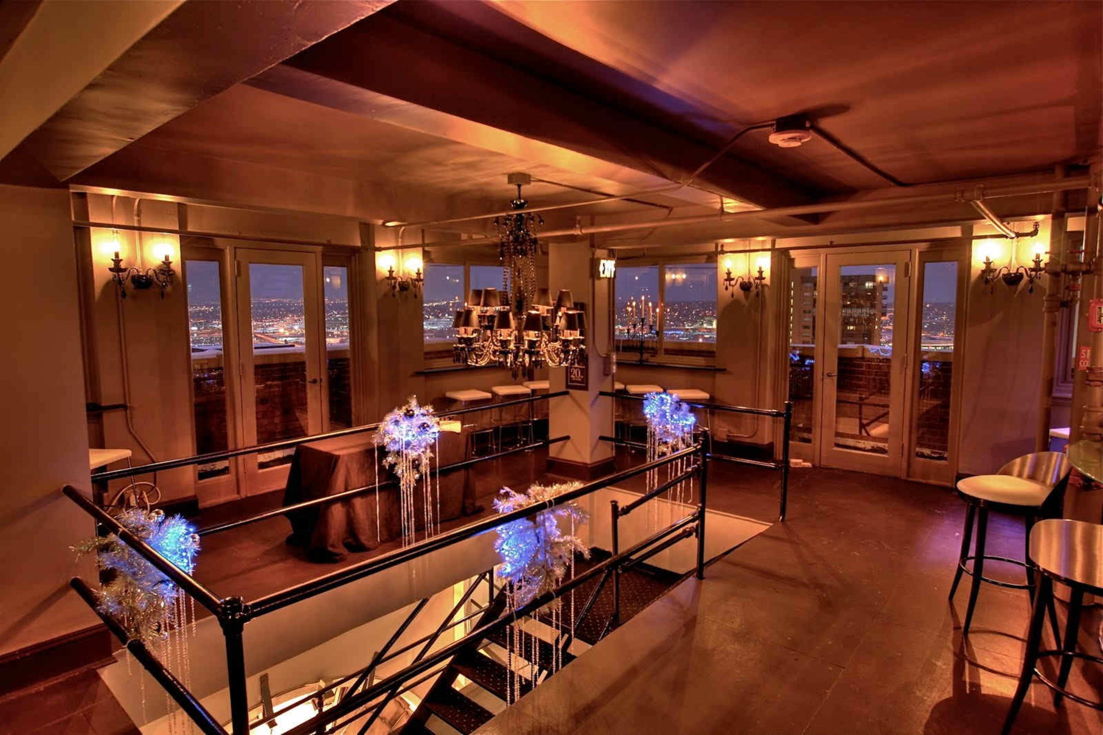 An elegantly lit interior space with a staircase leading to an upper level, featuring chandeliers and a bar area overlooking a city skyline at night.