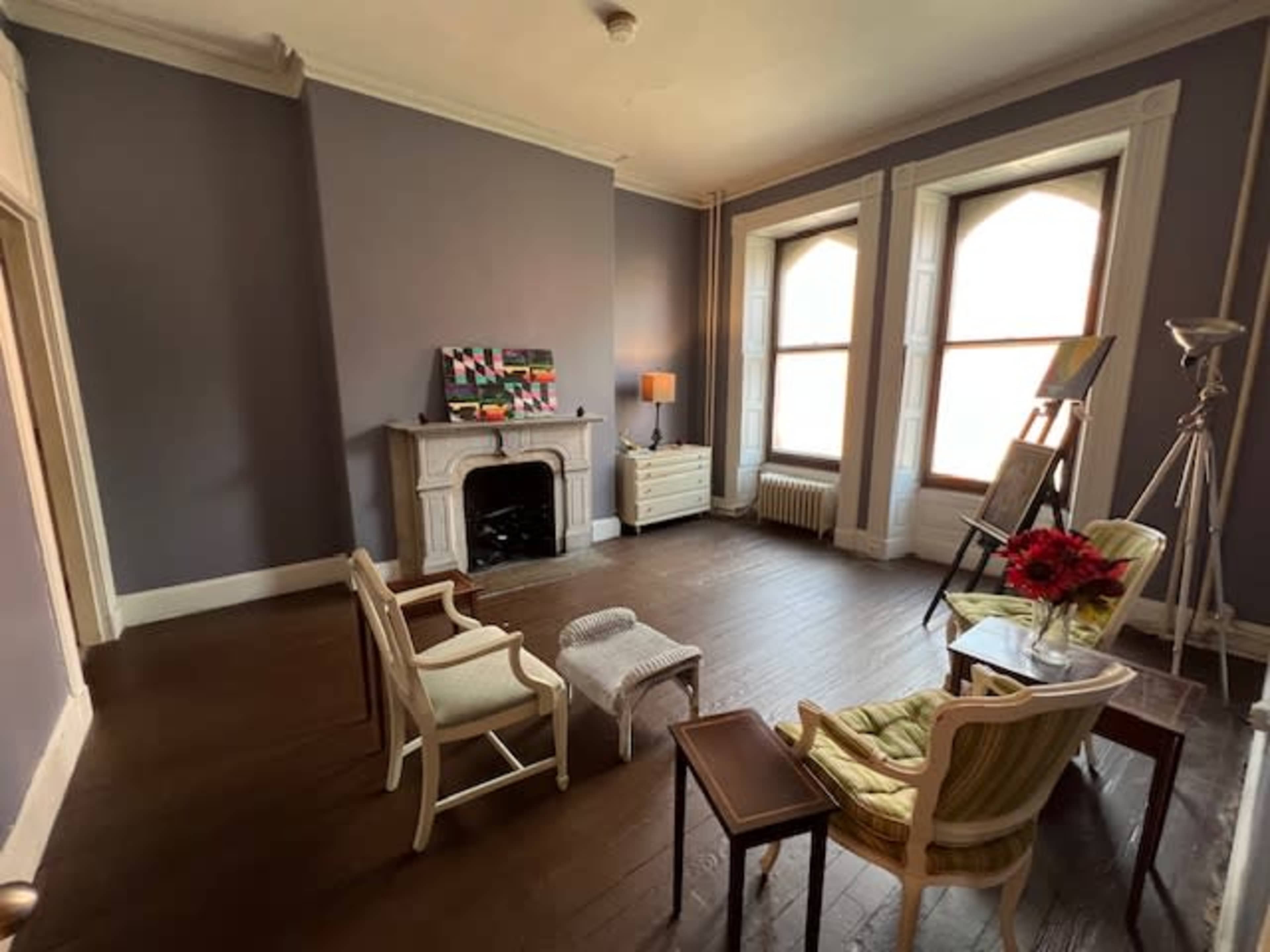 A spacious, empty living room features two armchairs, a small table, and large windows, with a decorative fireplace and a painting on the wall.