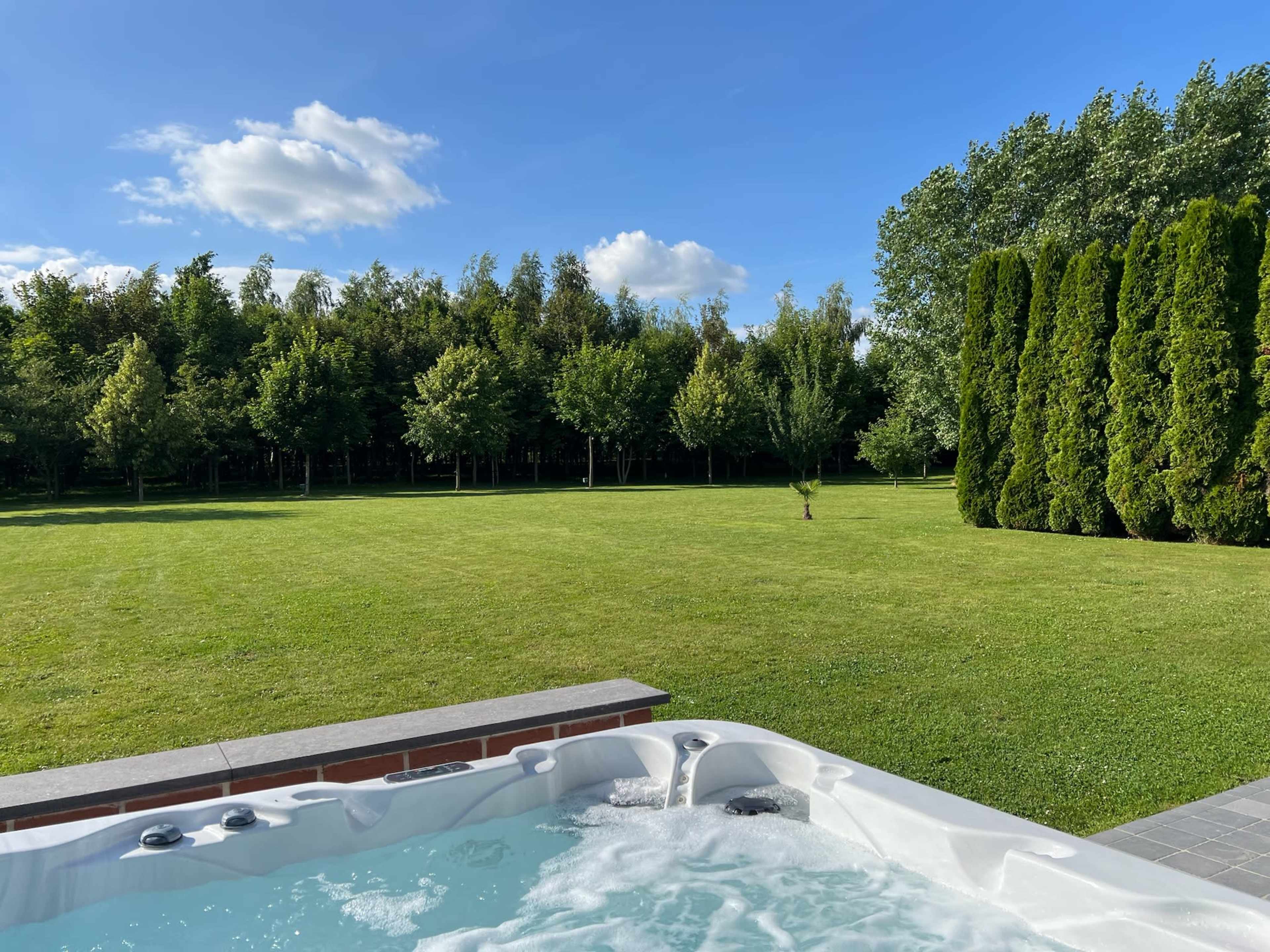 A hot tub sits at the edge of a manicured lawn, with a backdrop of trees and a clear blue sky.