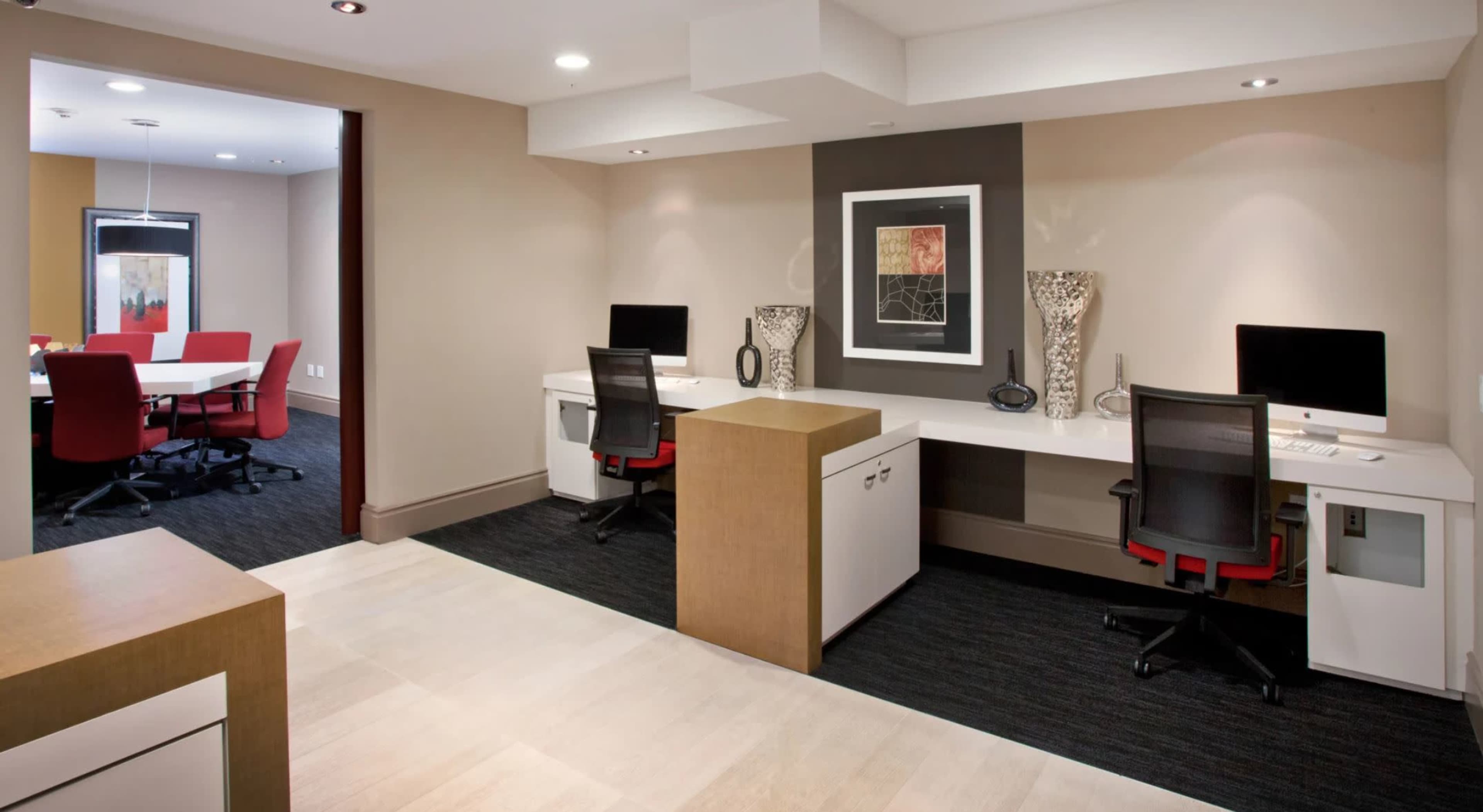 The image shows a modern office space with two desks facing each other, each equipped with a computer, alongside a meeting room with red chairs in the background.