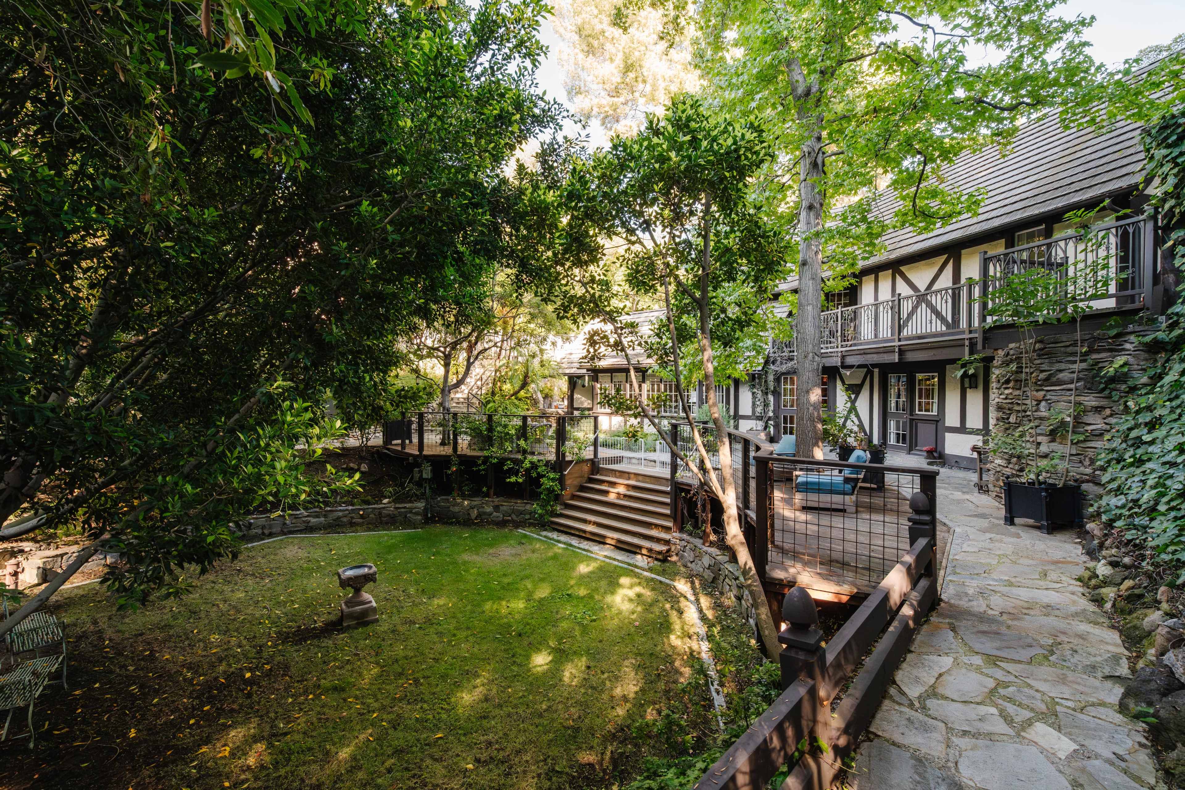 A stone pathway leads through a landscaped garden with a multi-level deck and a two-story house framed by trees.