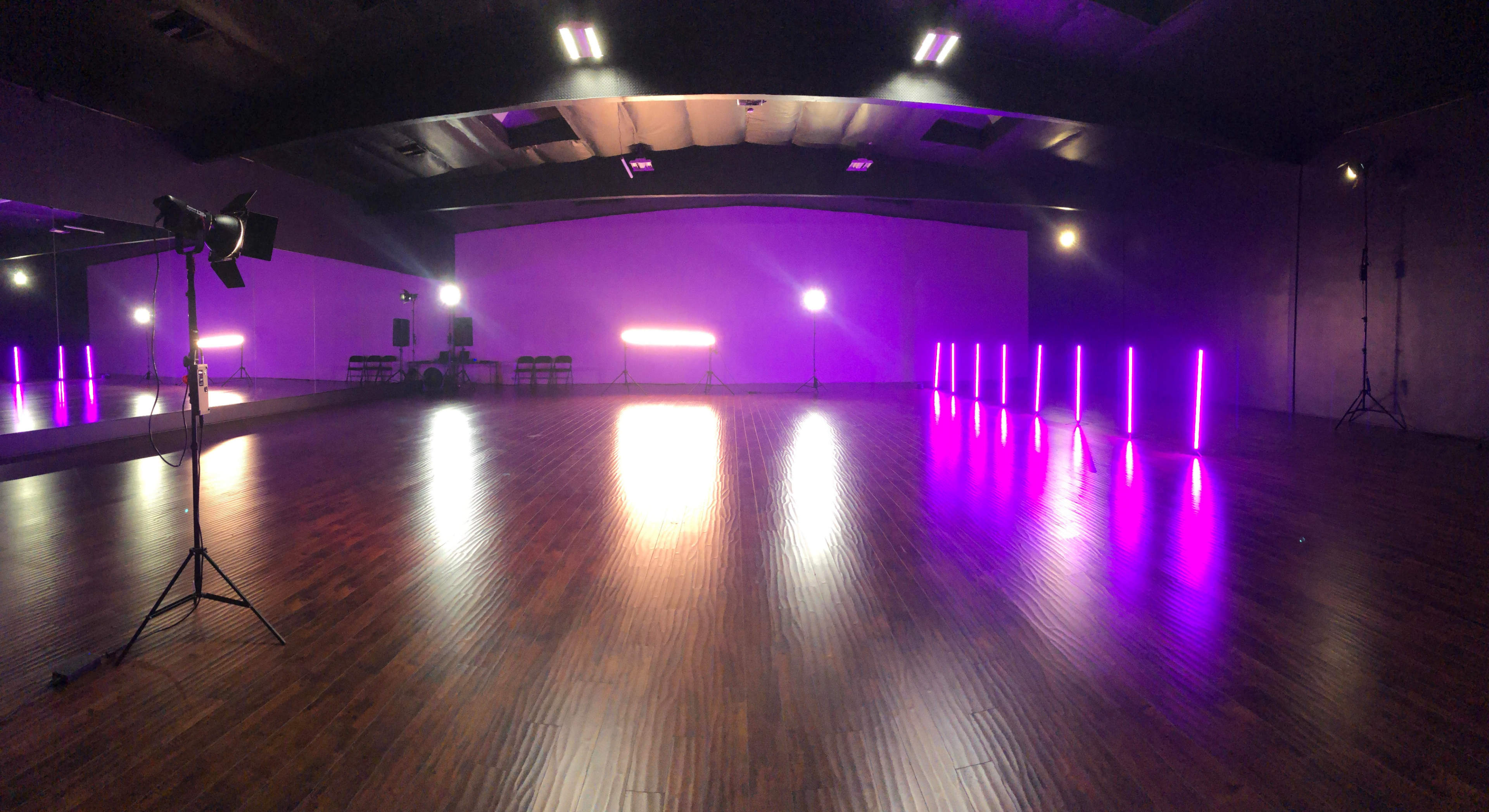 A spacious dance studio features polished wooden floors, illuminated by purple LED lights and various studio equipment.