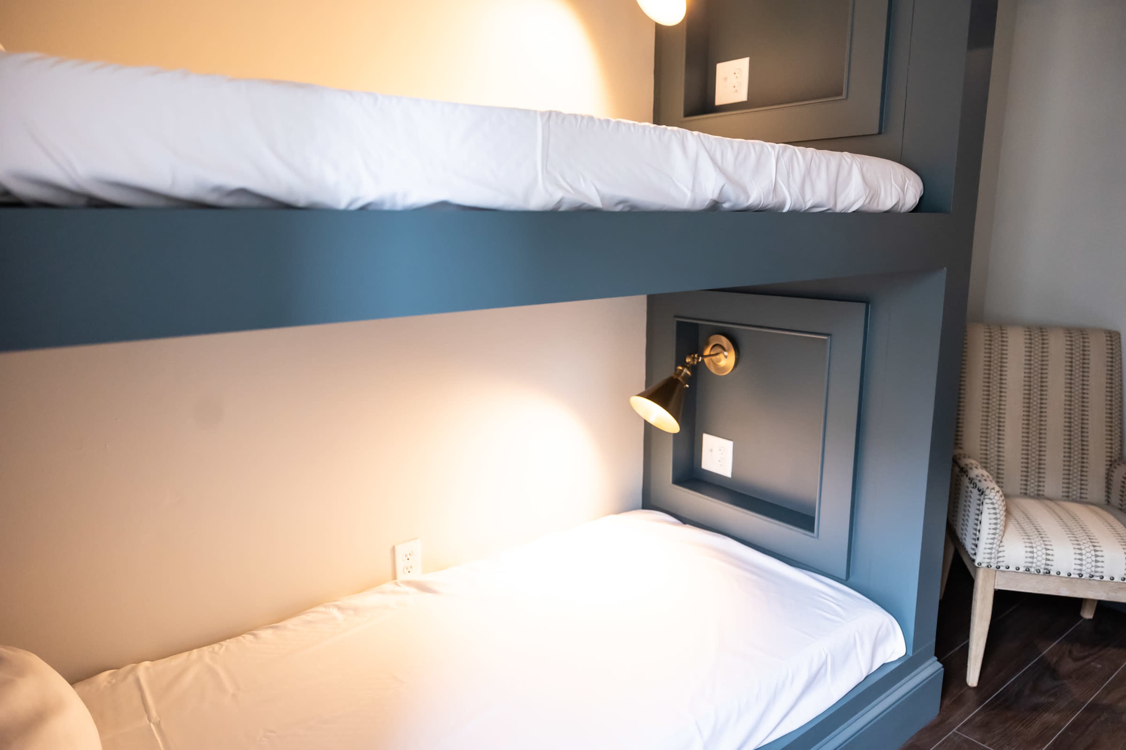 The image shows a compact room with a double-decker bunk bed, featuring a top and bottom sleeping area, along with a wall-mounted reading light and an armchair nearby.