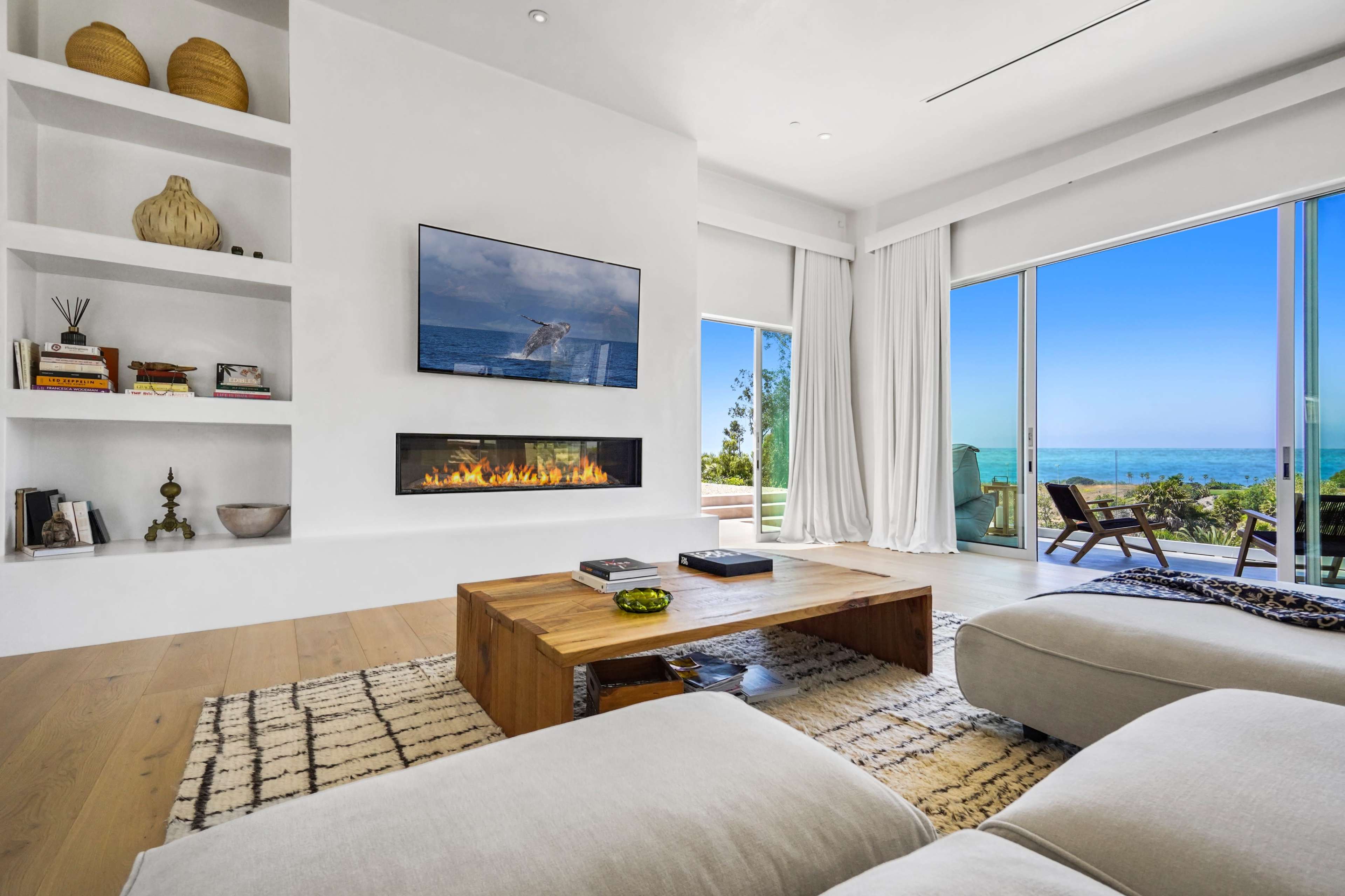 A modern living room features a wall-mounted television, a linear fireplace, and large windows that open to a view of the ocean.
