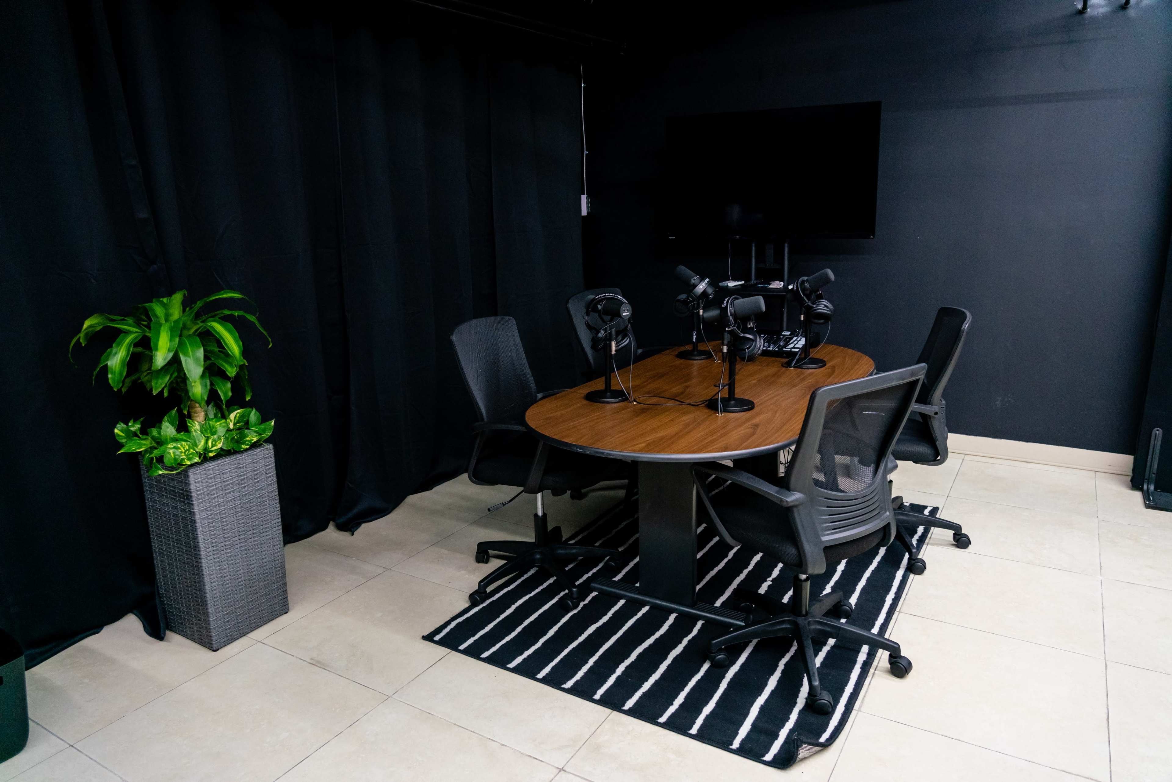 A modern conference room features a round wooden table with four chairs, microphones, a television on the wall, and a plant in a pot.