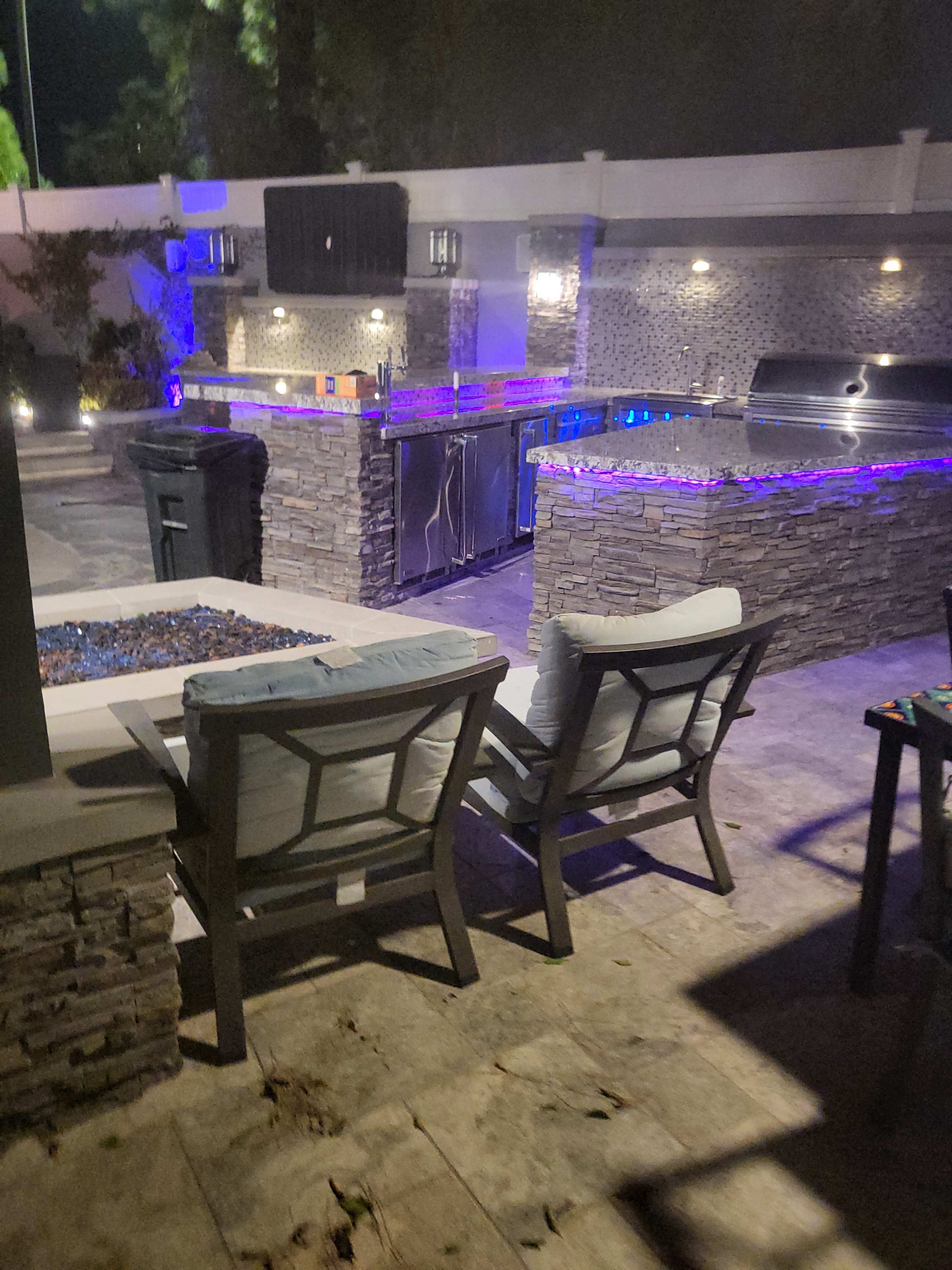 A modern outdoor kitchen features stone countertops, a grill, and ambient lighting, with two chairs positioned nearby.