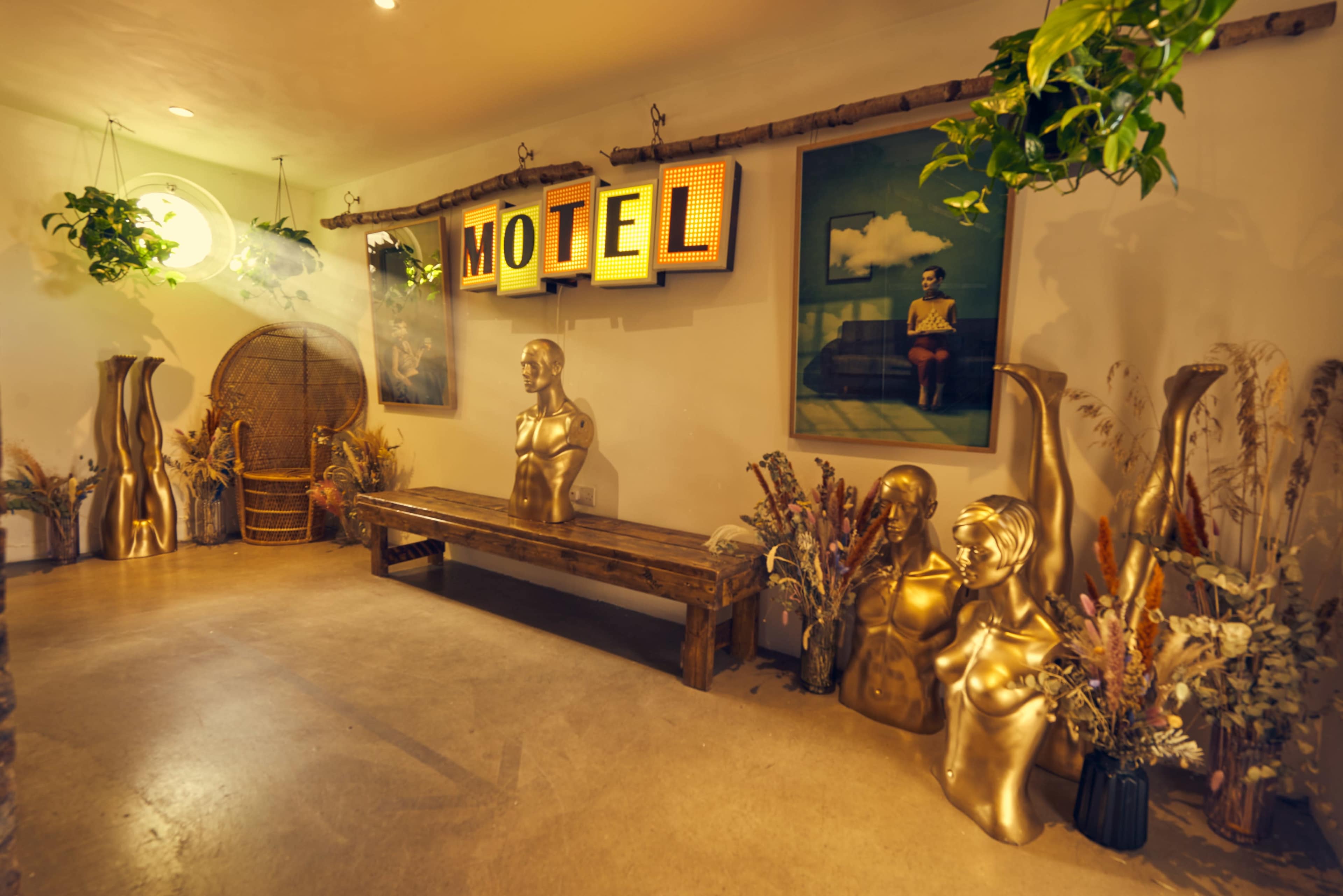 The interior of a stylish motel features two gold mannequins, a wooden bench, and framed art, all surrounded by decorative plants.