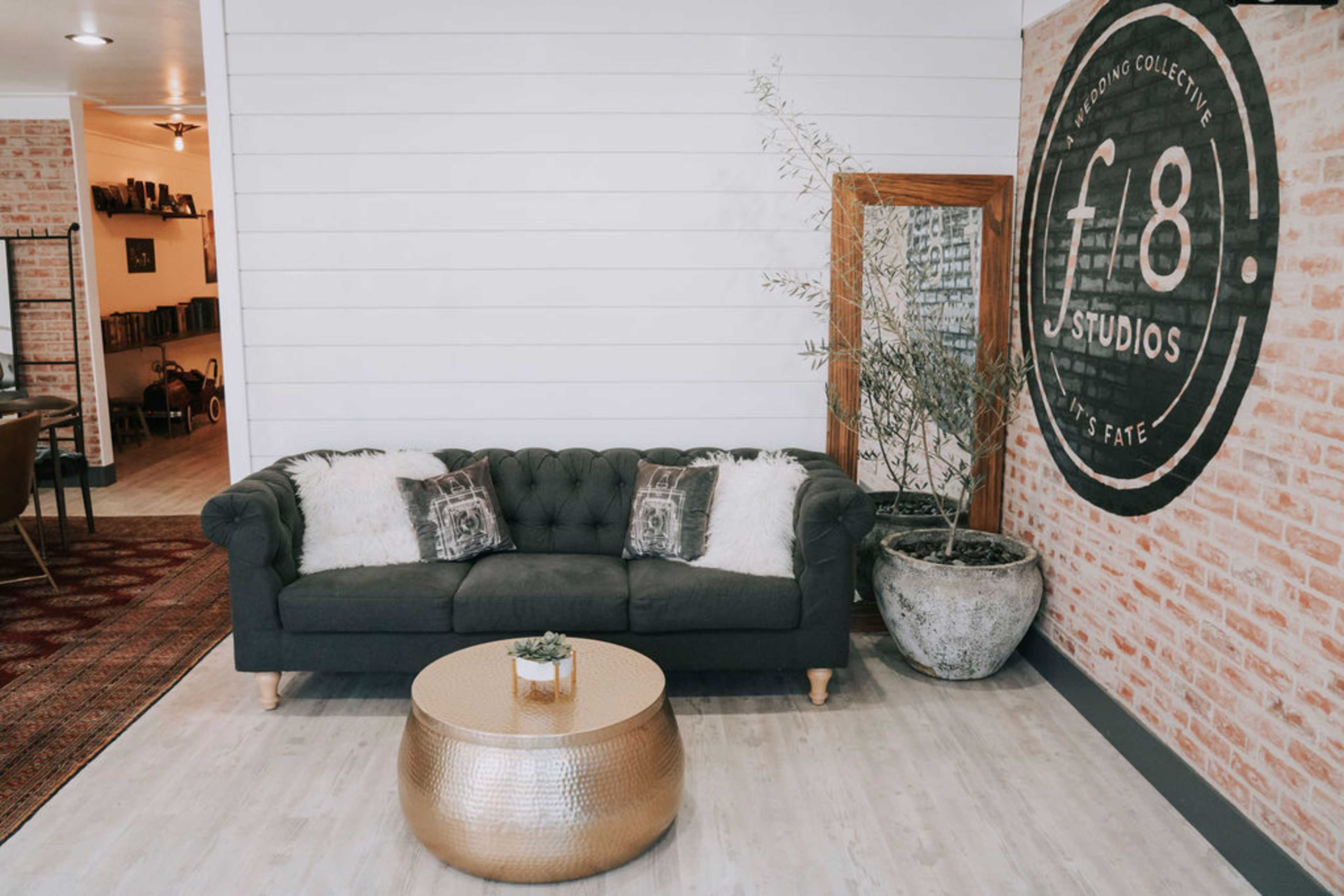A tufted dark sofa with decorative pillows, a round golden coffee table, and a large mirror against a brick wall in a modern interior space.