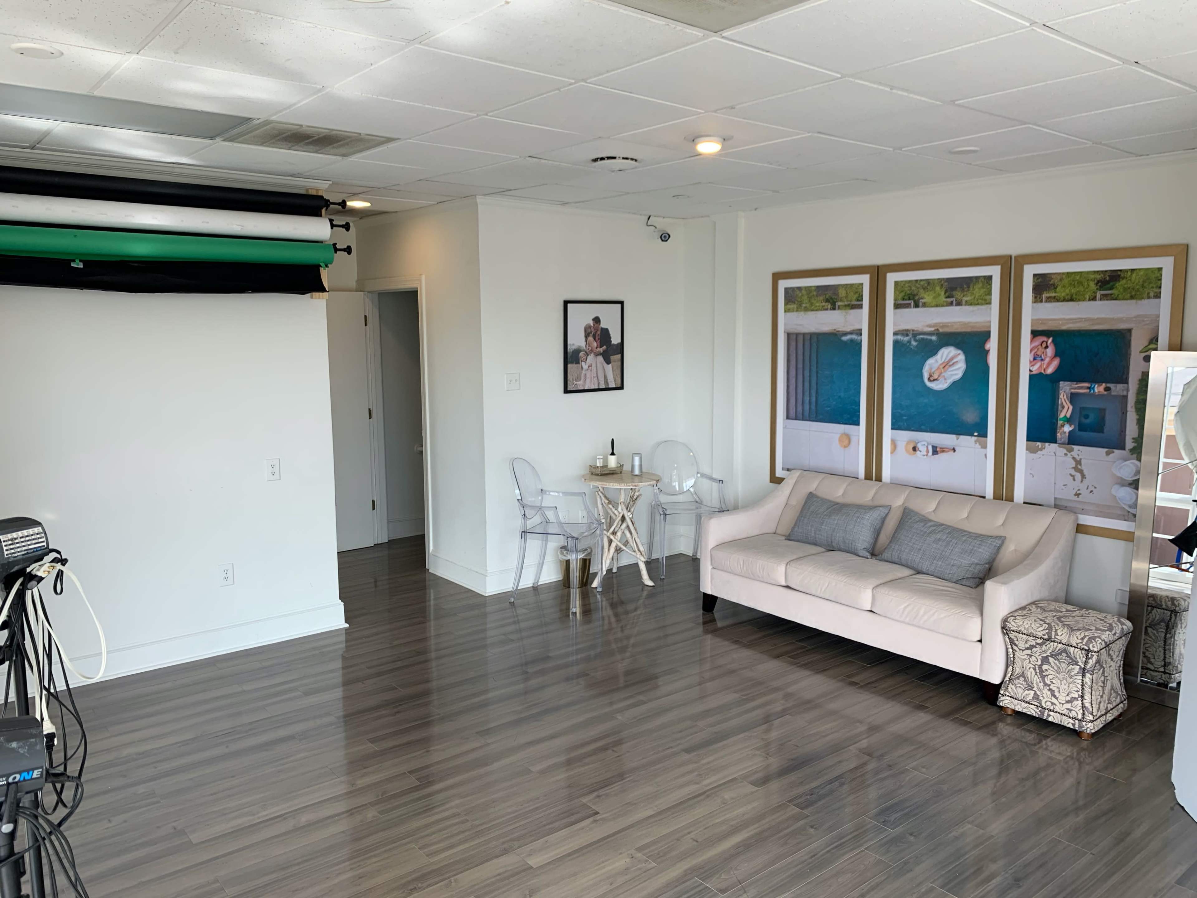 The image shows a bright, minimalist studio space featuring a small dining table with chairs, a couch, and framed photographs on the wall.