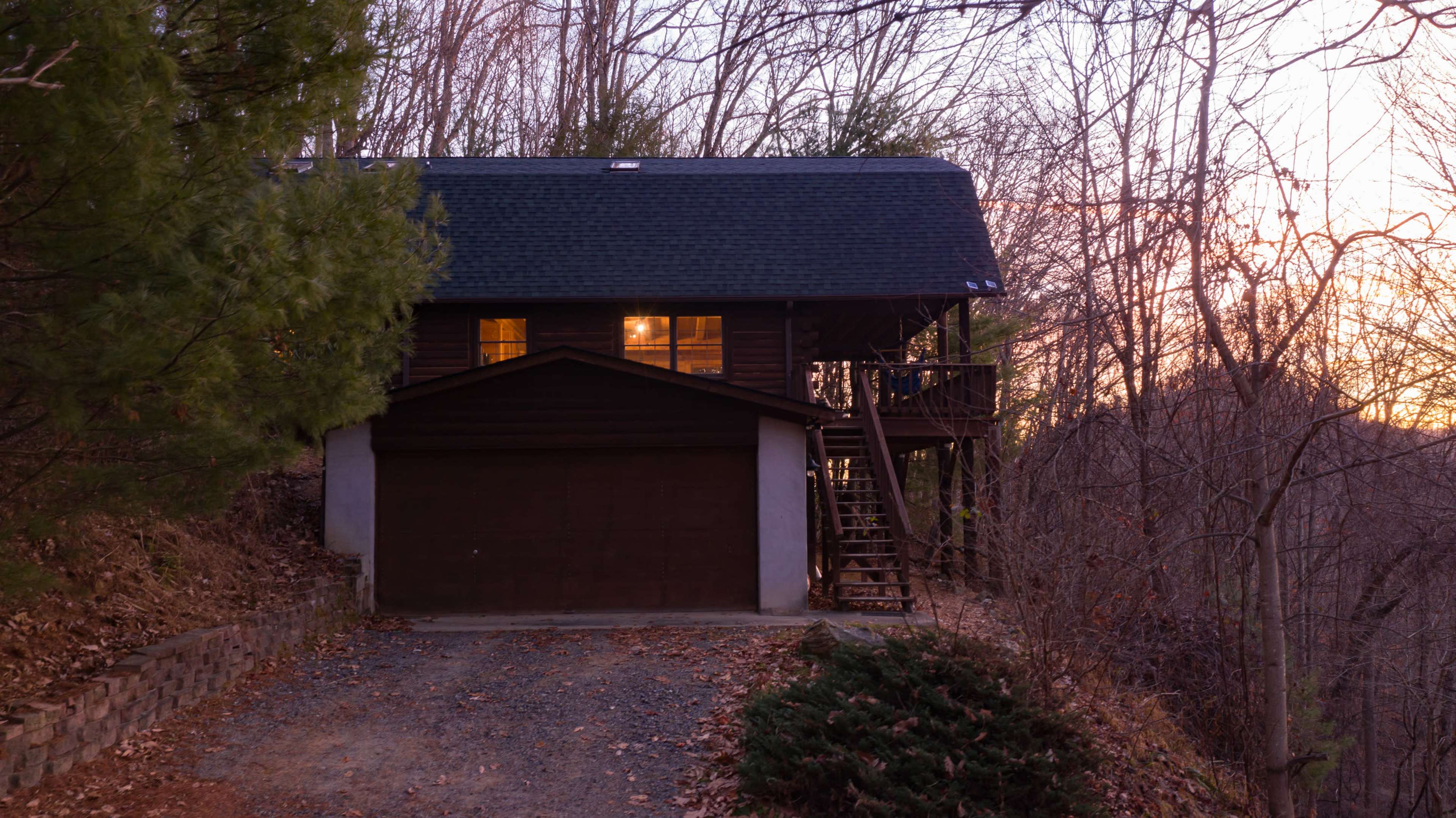 A wooden house with a garage sits on a gravel driveway surrounded by bare trees at sunset.