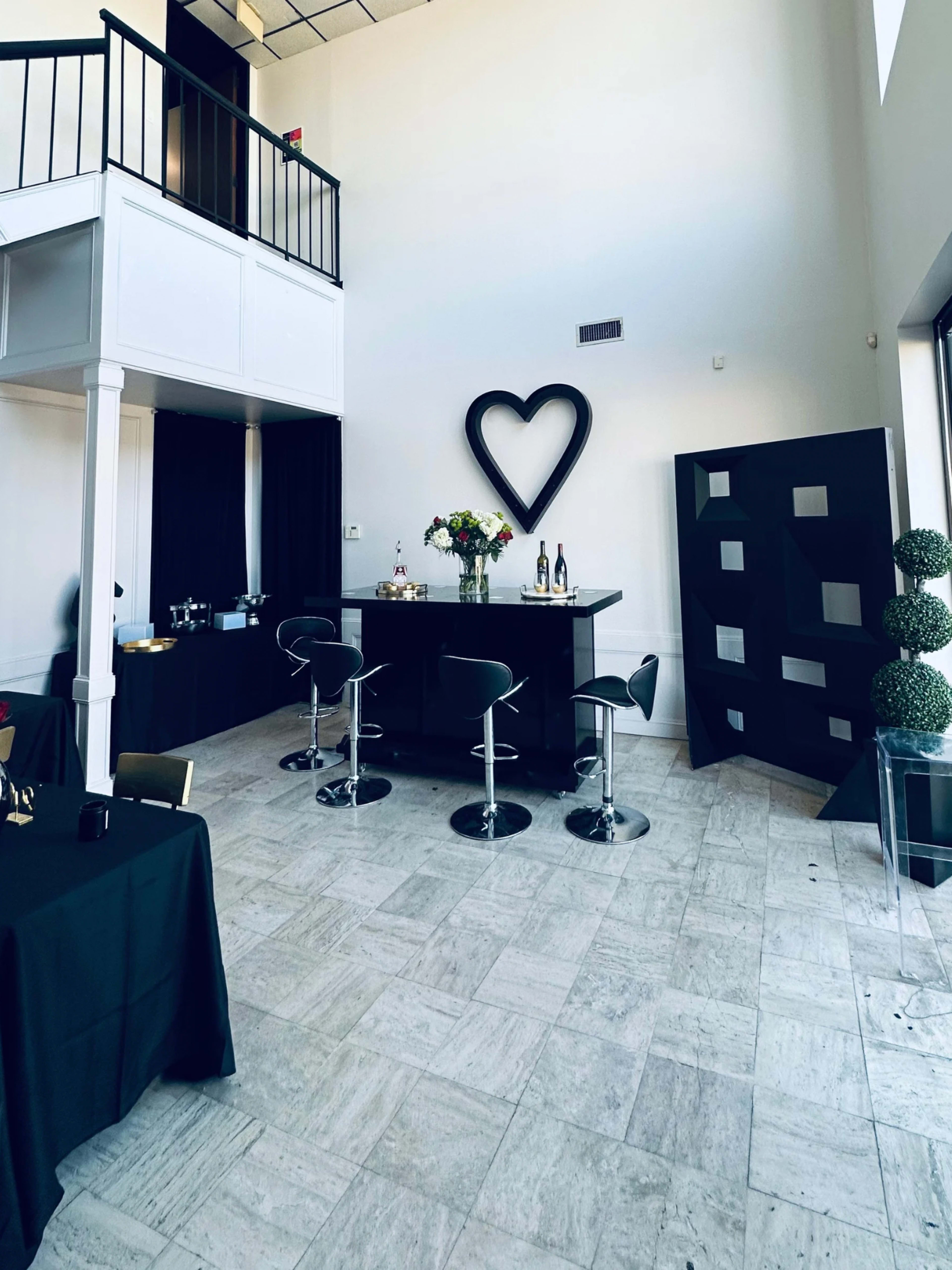 The image shows a spacious interior with a bar counter, black stools, a large heart-shaped decoration on the wall, and potted topiary plants.
