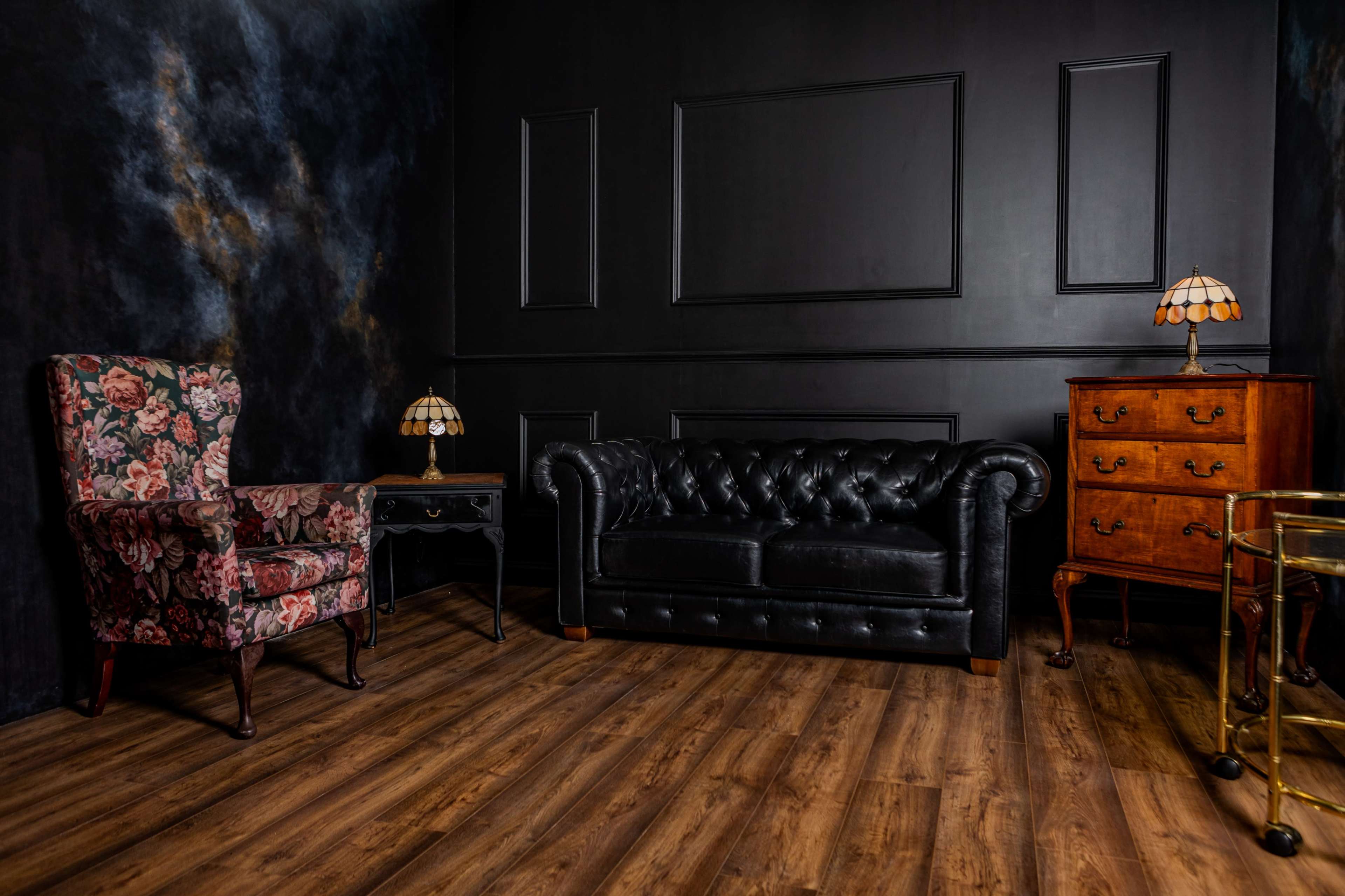 The image depicts a stylish interior featuring a black leather Chesterfield sofa, a floral patterned armchair, an ornate wooden side table, and two vintage lamps, all set against a darkly painted wall with textured accents.