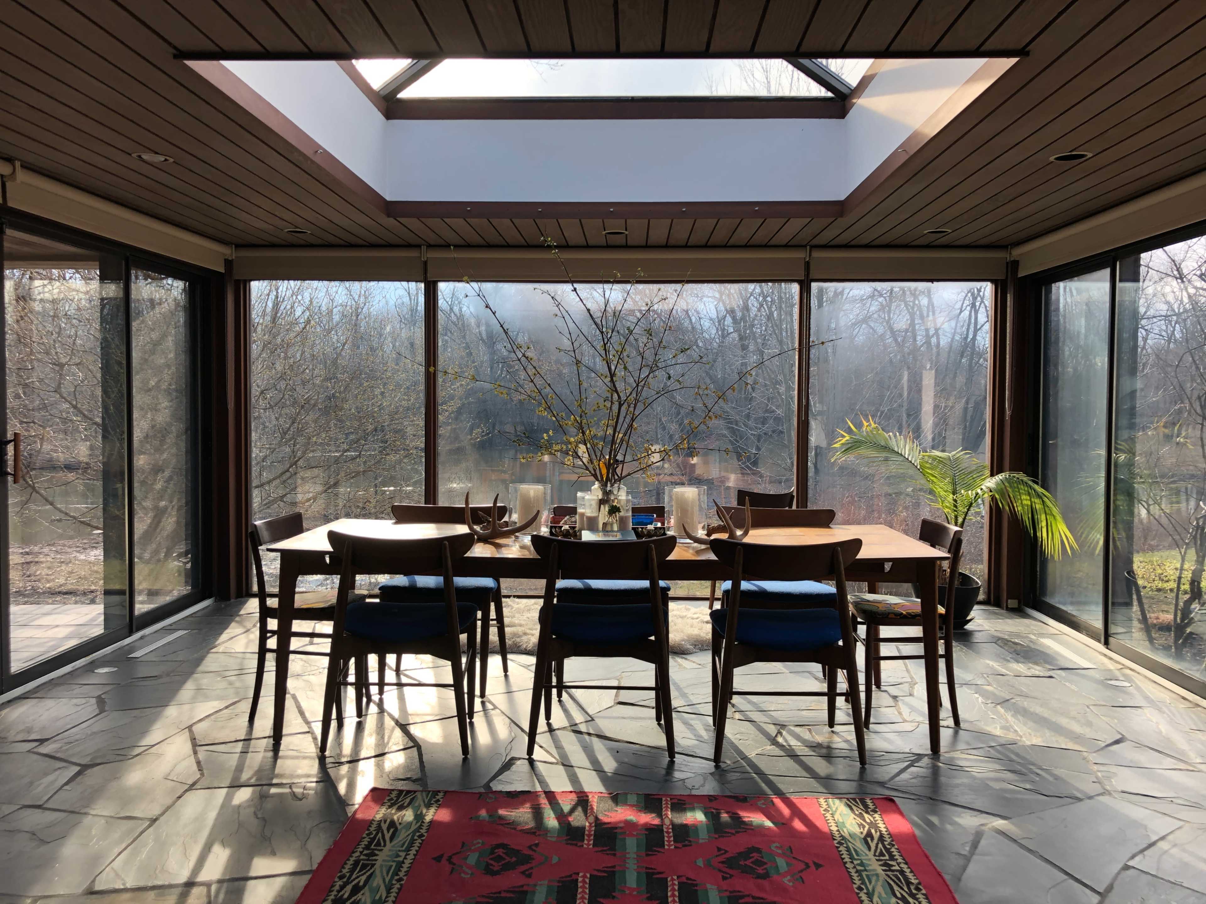 A dining area with a large table surrounded by chairs overlooks a serene outdoor landscape through expansive glass doors and a skylight.