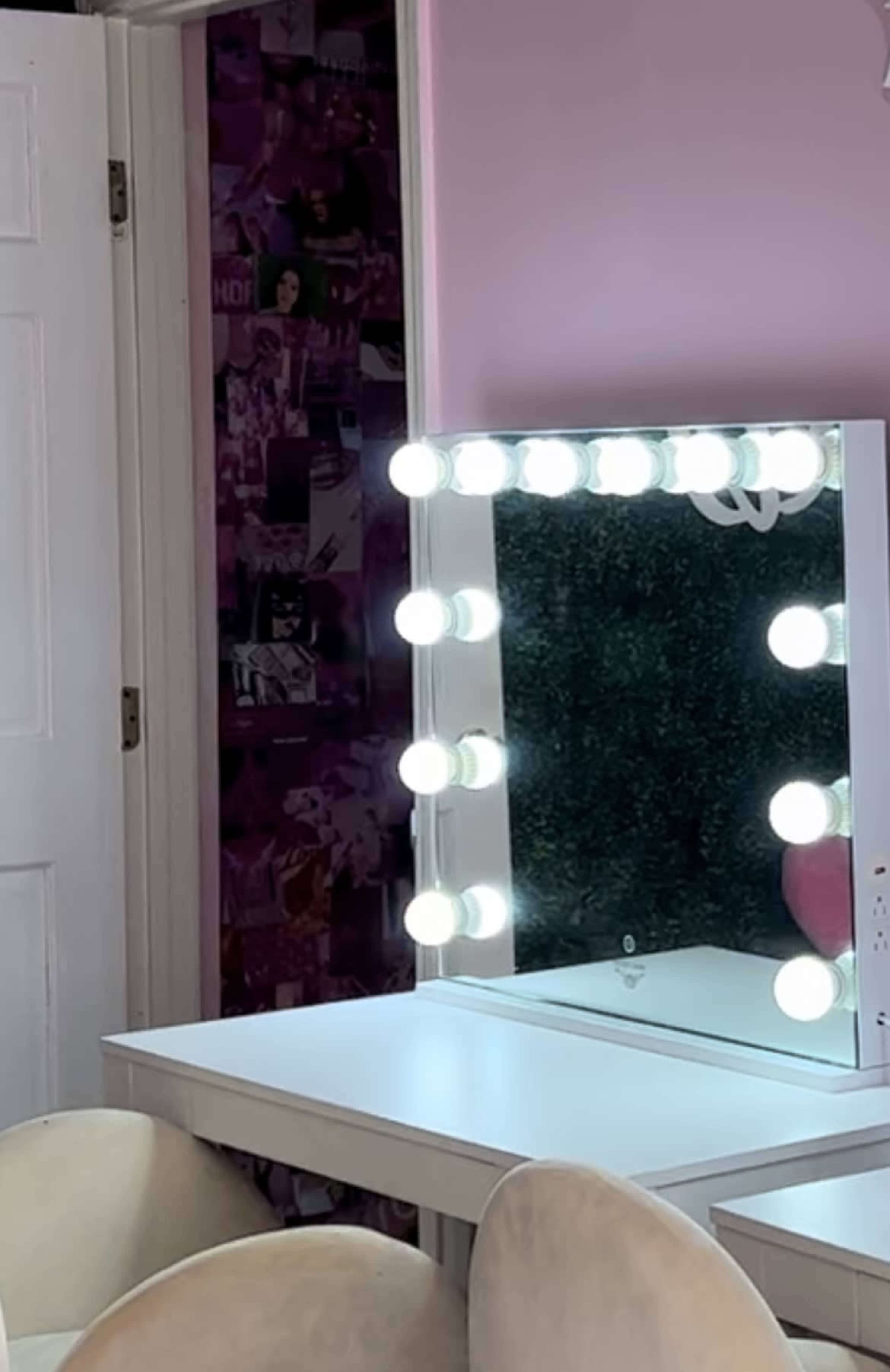 A vanity mirror with bright light bulbs is positioned against a pink wall, next to a white table and a door in the background.