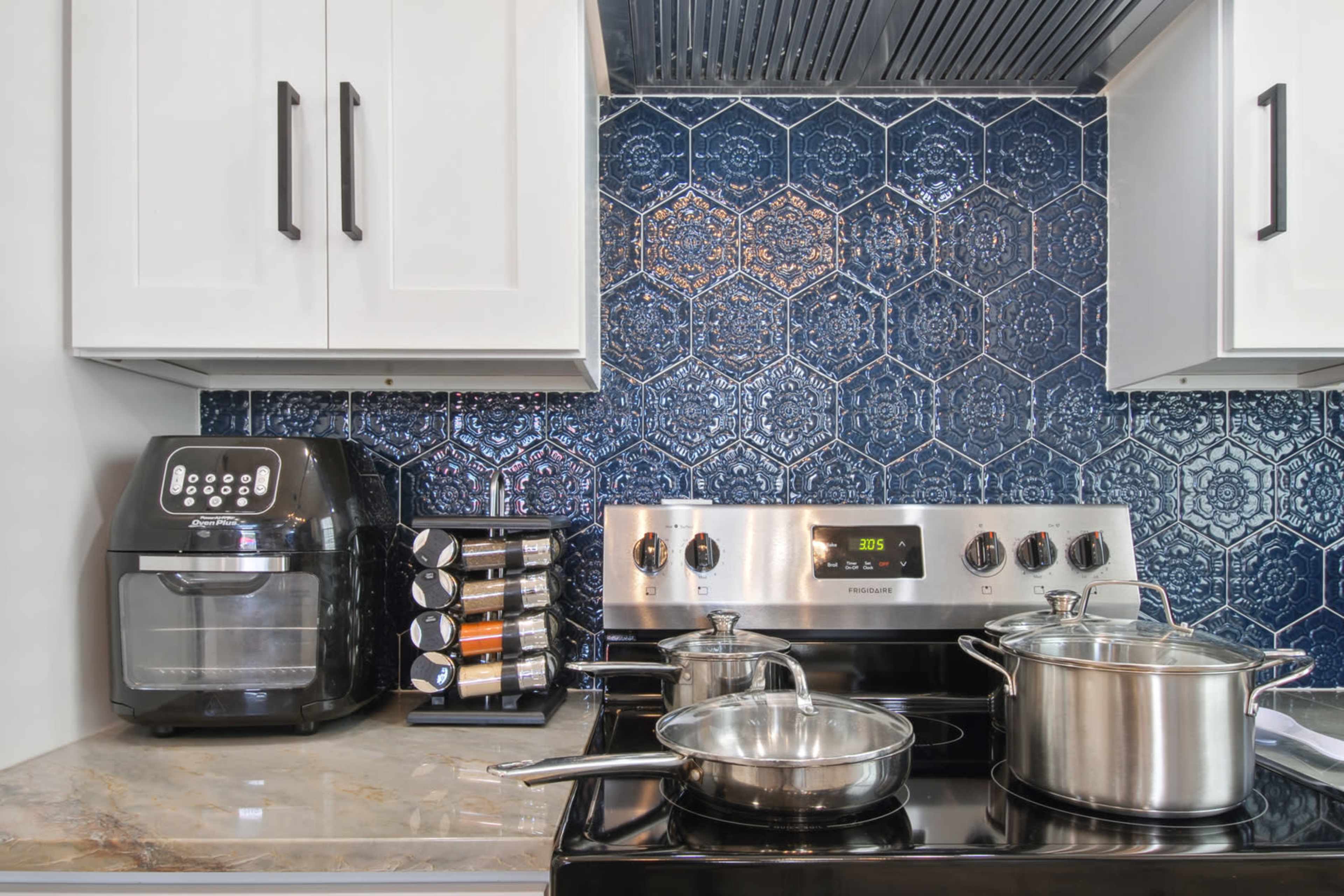 The image shows a modern kitchen with a stainless steel stove, pots on the burner, an air fryer on the counter, and a spice rack beside a blue tiled backsplash.