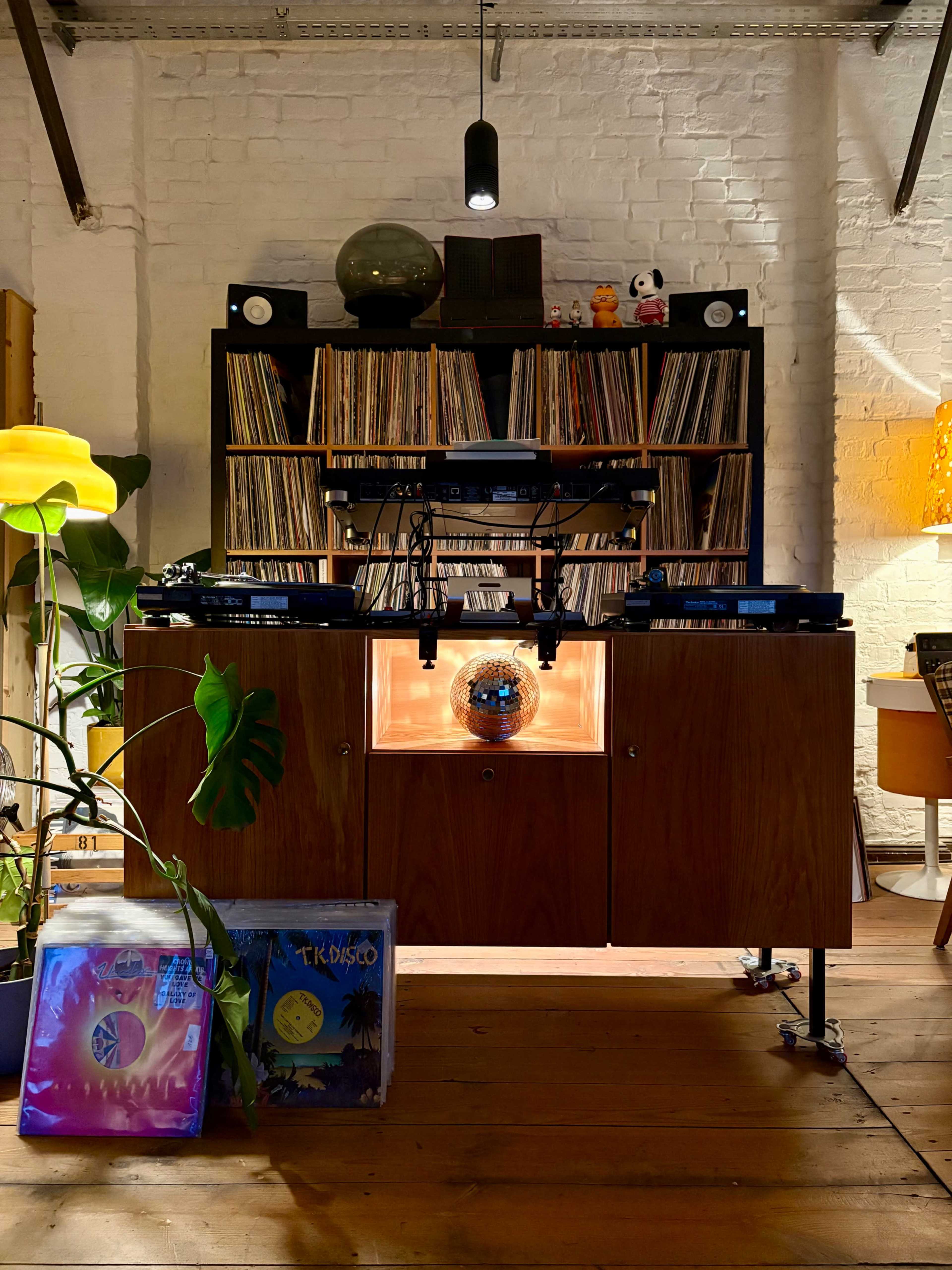 A wooden console with a record player and disco ball is positioned in front of a wall of vinyl records, surrounded by plants and colorful decor.