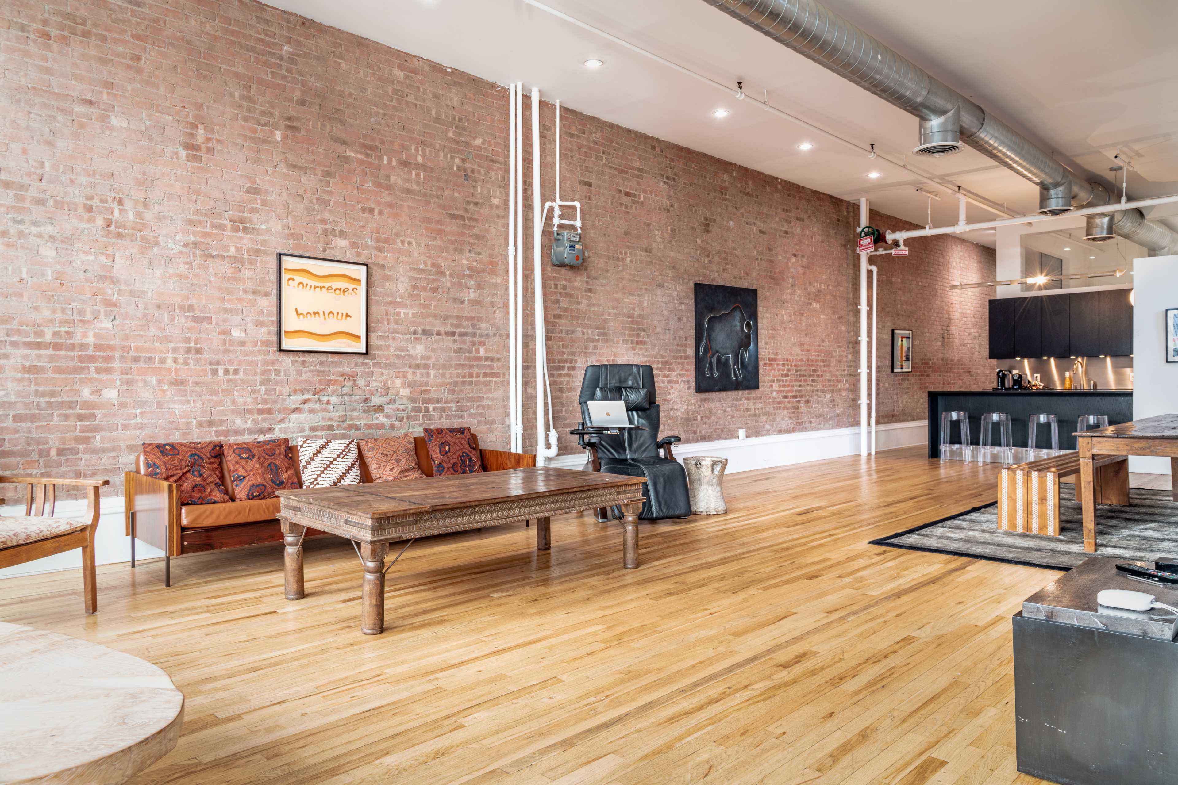 The image shows a spacious loft with exposed brick walls, wooden flooring, and a mix of contemporary and rustic furniture, including a couch, a coffee table, and a black kitchenette in the background.