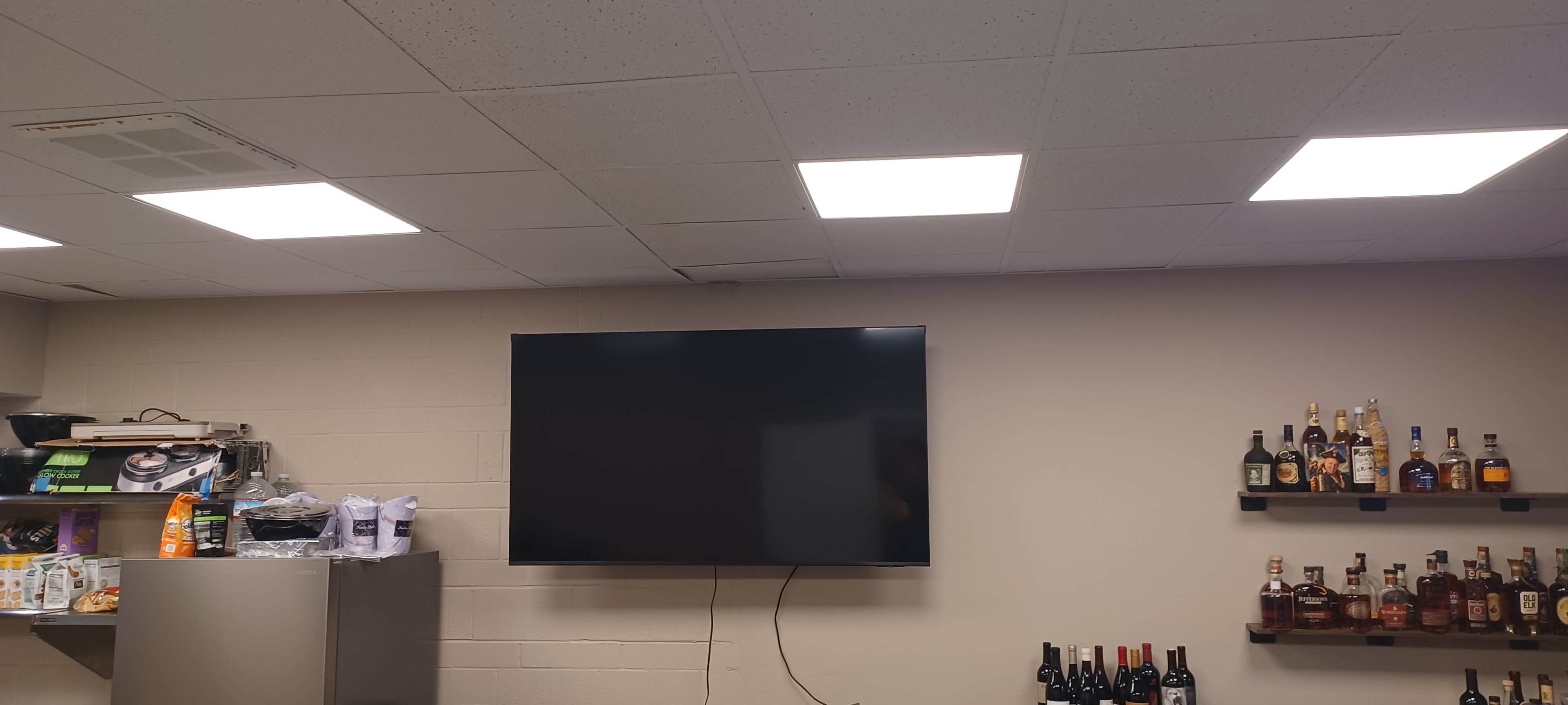 The image shows a room with a wall-mounted television, overhead square lights, and shelves displaying various bottles.