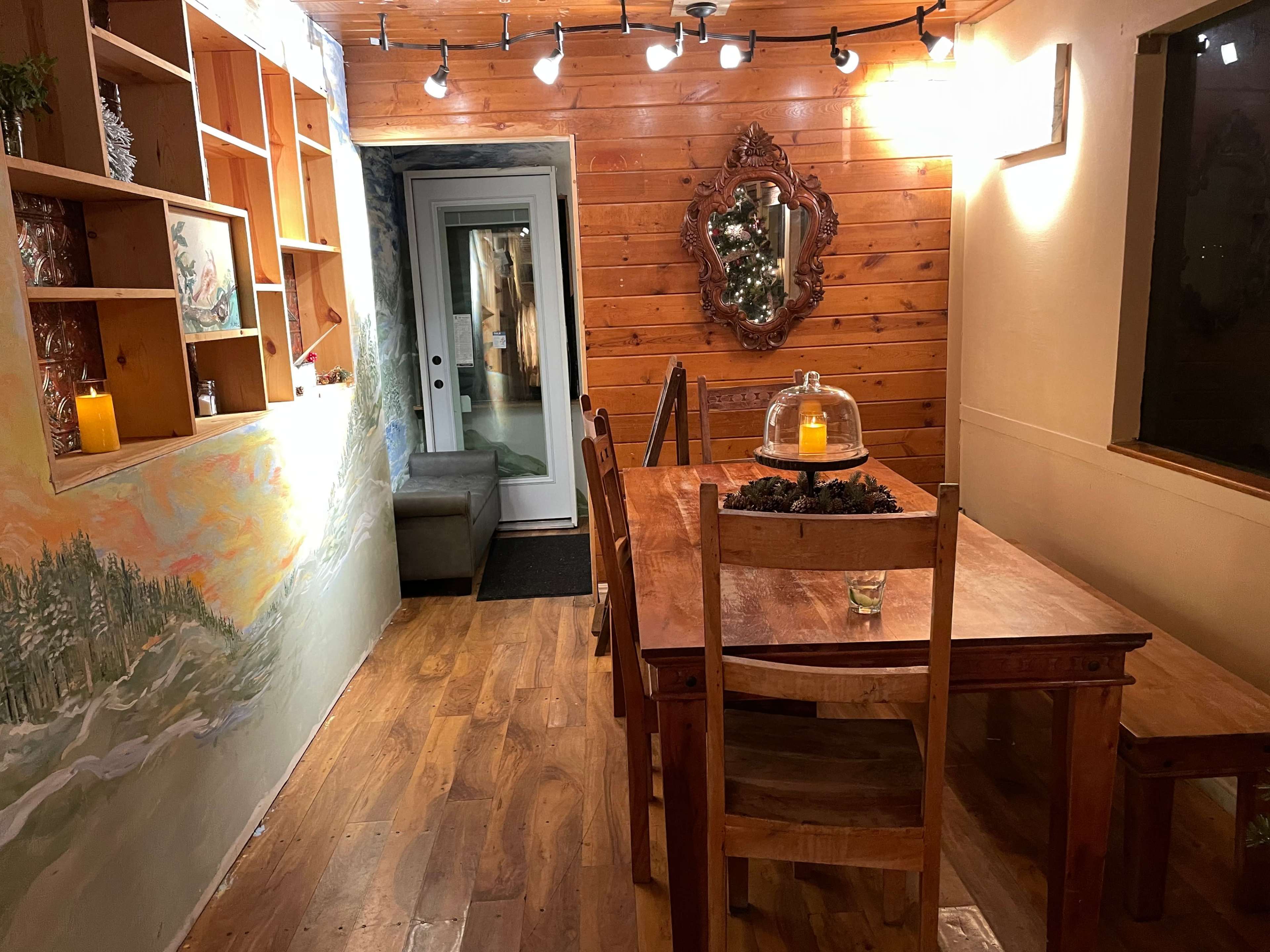 The image shows a cozy dining area with a wooden table and chairs, illuminated by warm lights, featuring a wall painted with a landscape and a rustic mirror.