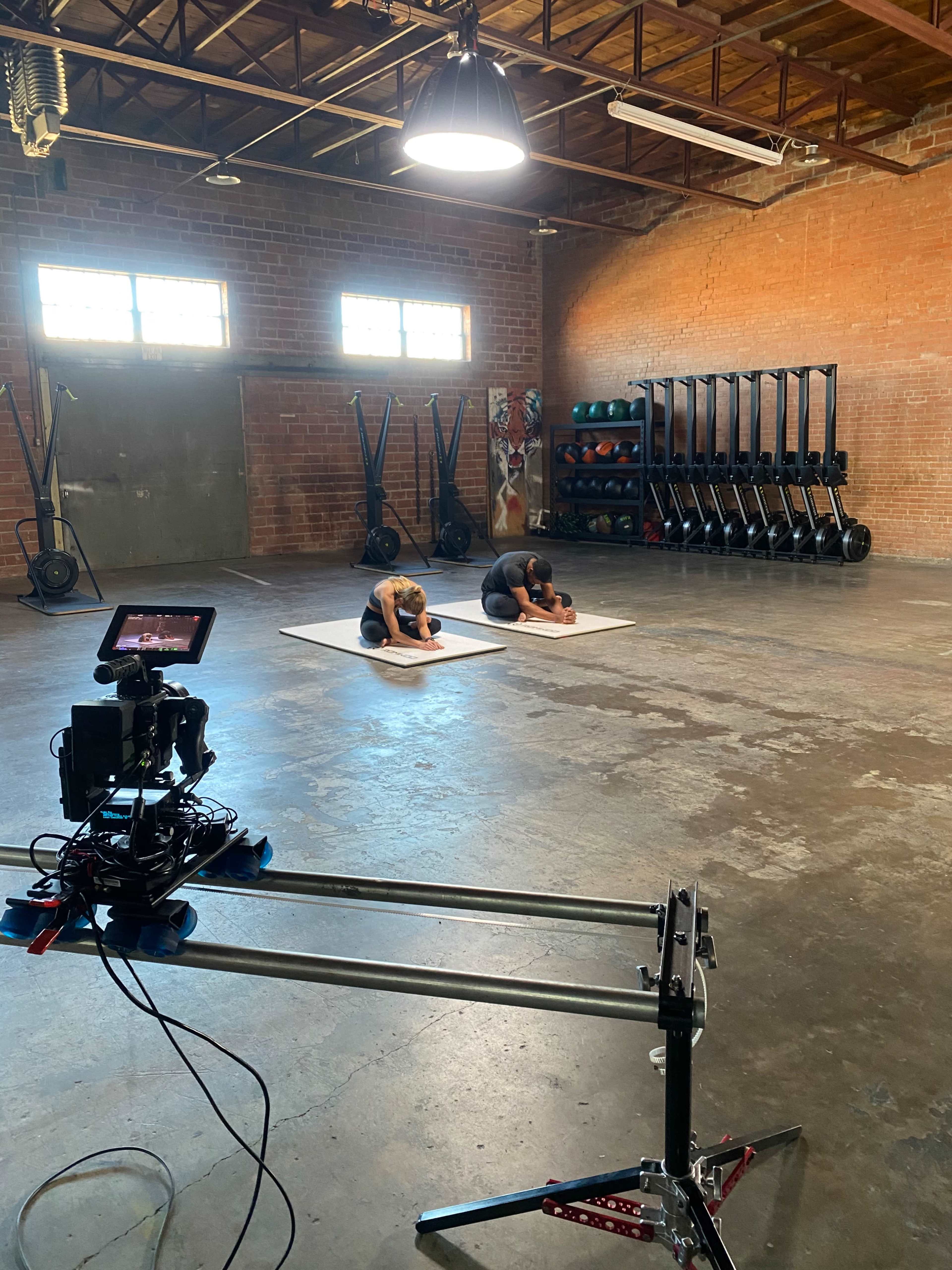 Two individuals are performing exercises on yoga mats in a spacious gym with various fitness equipment in the background.