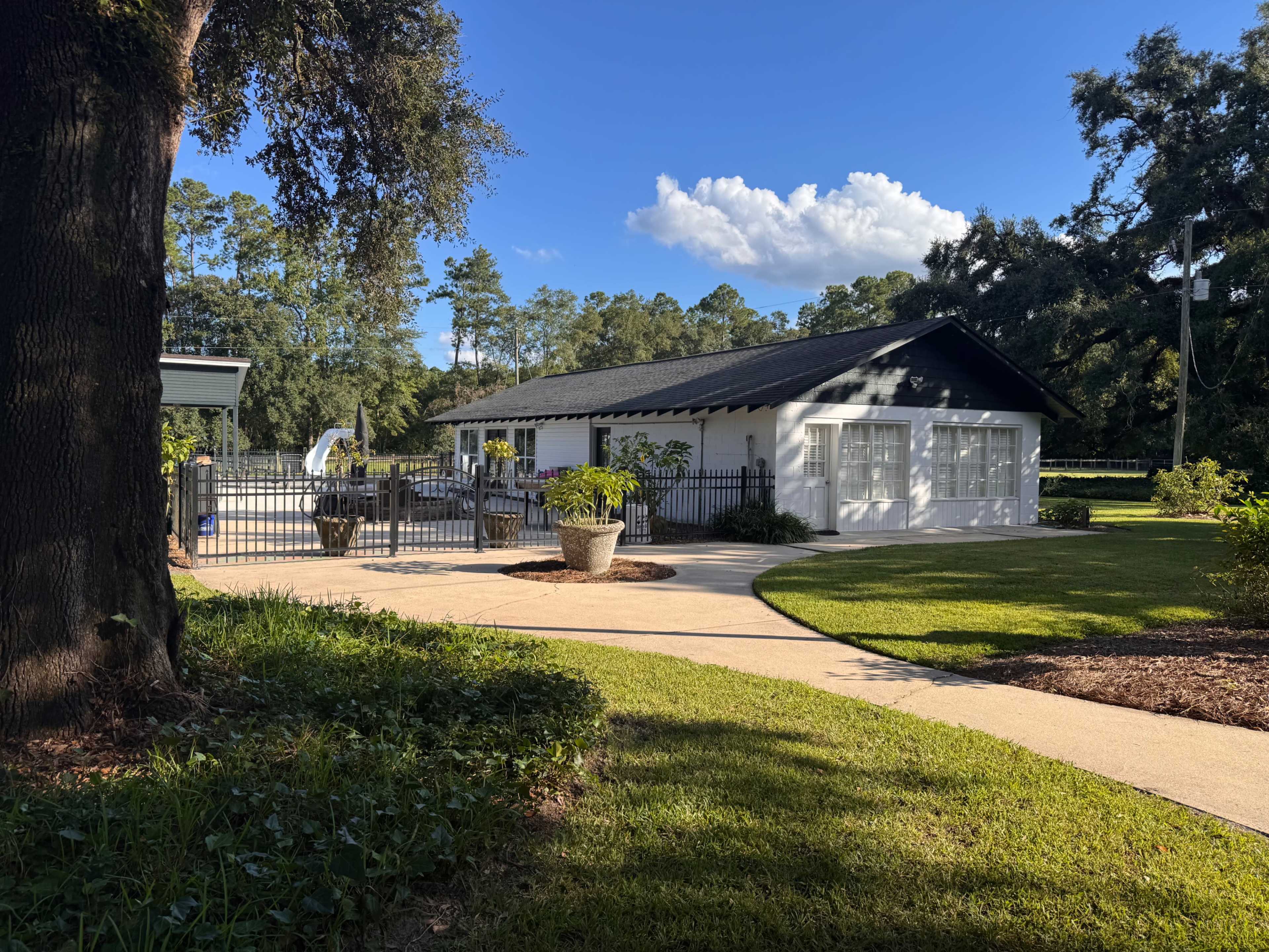 A white building with a black roof sits behind a gate, surrounded by a well-manicured lawn and trees.